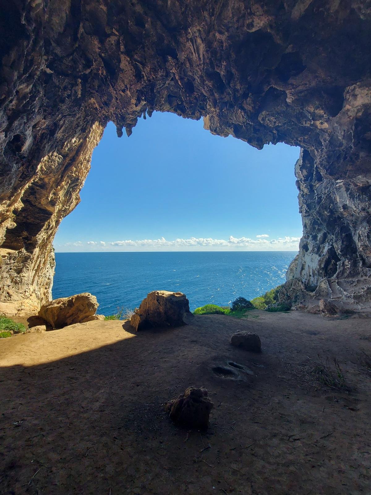 View from inside a cave looking out towards the ocean, with blue sky and some clouds. The cave has rocky walls and floor with a few large rocks inside.