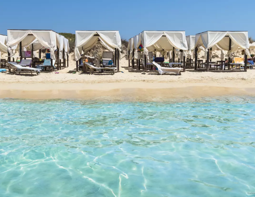 Beach with cabanas and chairs facing the ocean, sunny weather, clear blue water in the foreground.