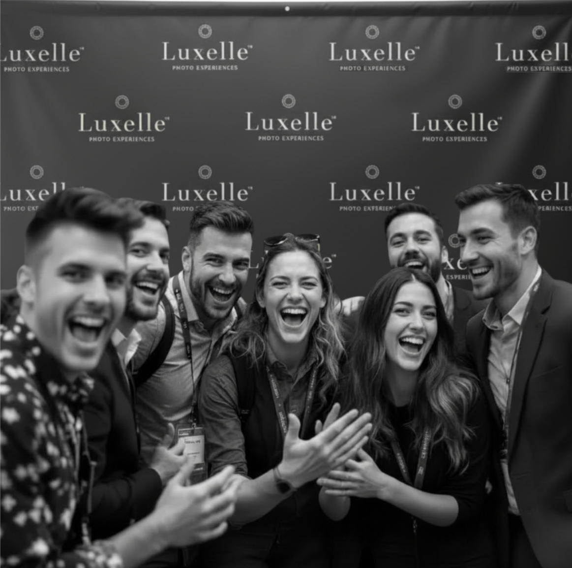 Group of seven smiling people at the Luxelle photo experiences event, posing together with a branded backdrop in front of a Photo booth