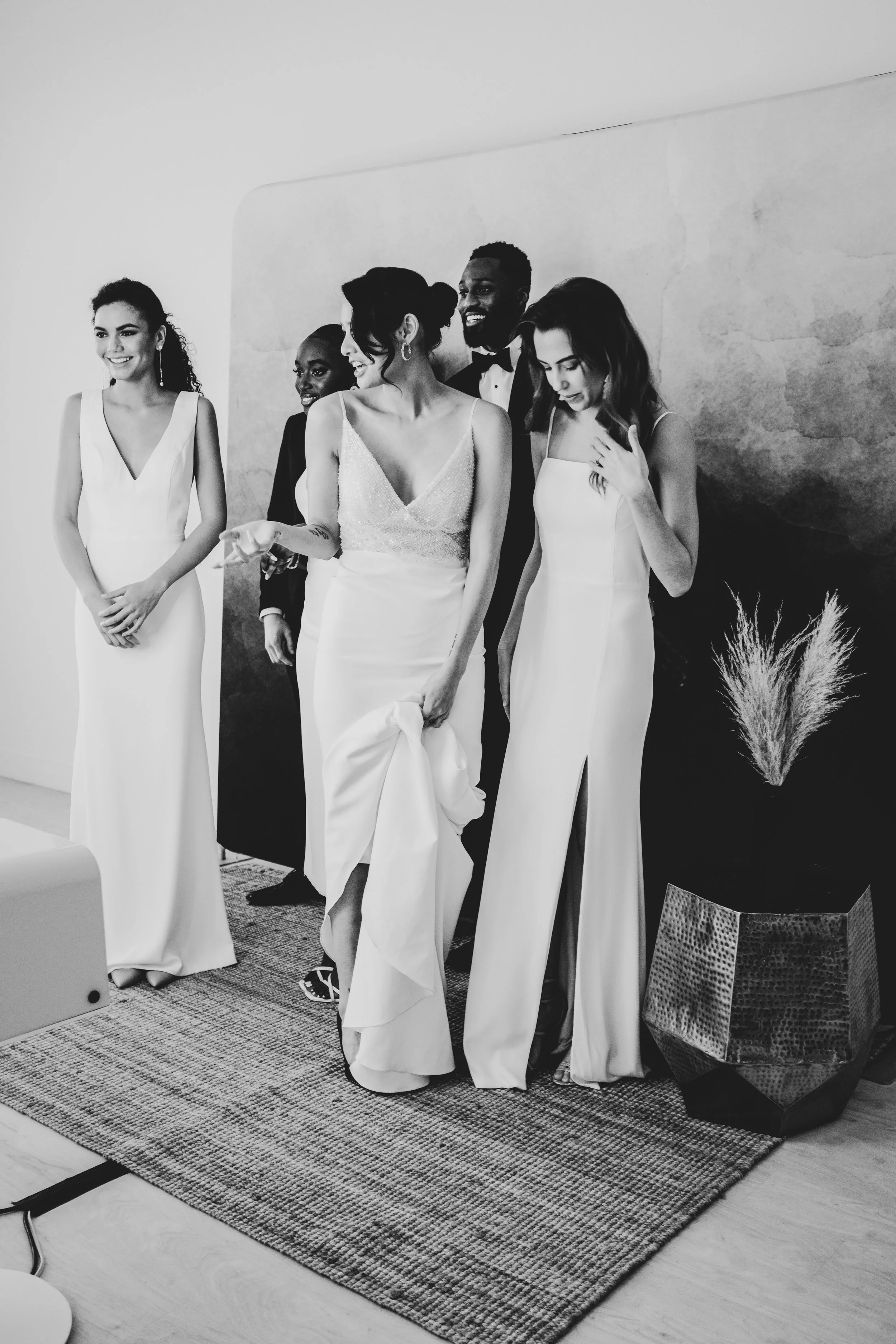 Group of six people at a wedding or formal event, taking selfie pictures behind a photobooth camera provided by Luxelle in front of a backdrop, with five women and one man, all dressed in elegant attire, standing on a patterned rug indoors.
