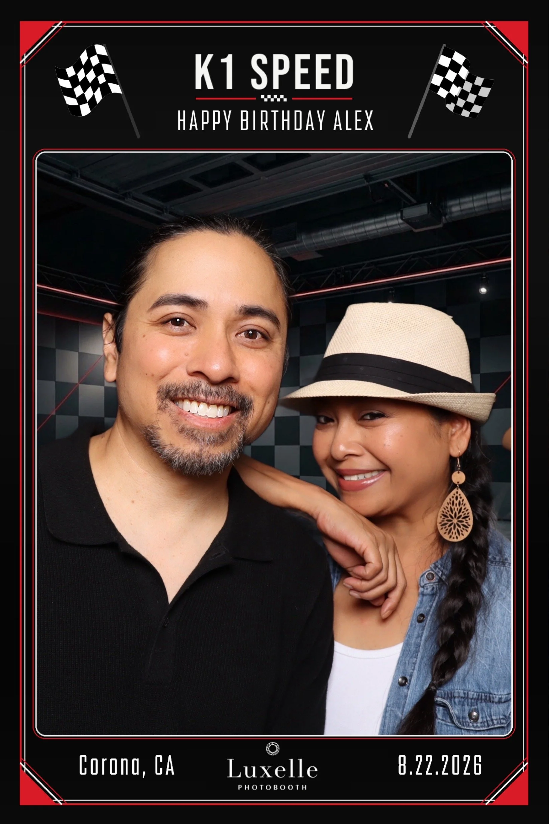 A photo of two smiling people, a man and a woman, posing together in a photo booth with a black and red border. The man has short dark hair, a mustache, and a beard, and is wearing a black shirt. The woman has long dark hair in a braid, is wearing a hat, earrings, and a denim jacket. The border includes text indicating it's a themed birthday celebration for Alex, with flags, and it mentions the location as Corona, CA, the place as Luxelle Photobooth, and the date as 8.22.2026.