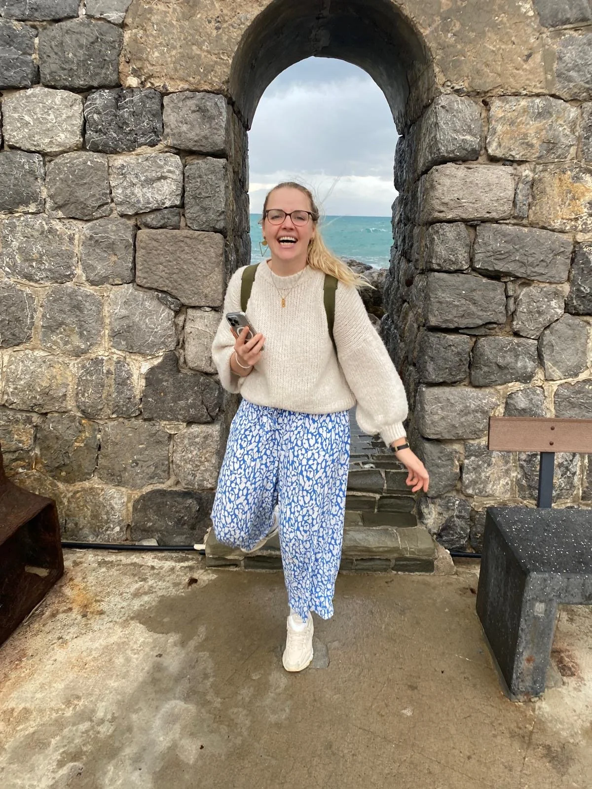 Junge Frau steht vor einer Steinemauer mit einem torförmigen Durchblick, im Hintergrund ist das Meer und ein stürmischer Himmel. Sie trägt eine weiße Pullover, blaue Hosen mit weißem Muster, weiße Schuhe, trägt eine Brille und hält ein Smartphone in der Hand.