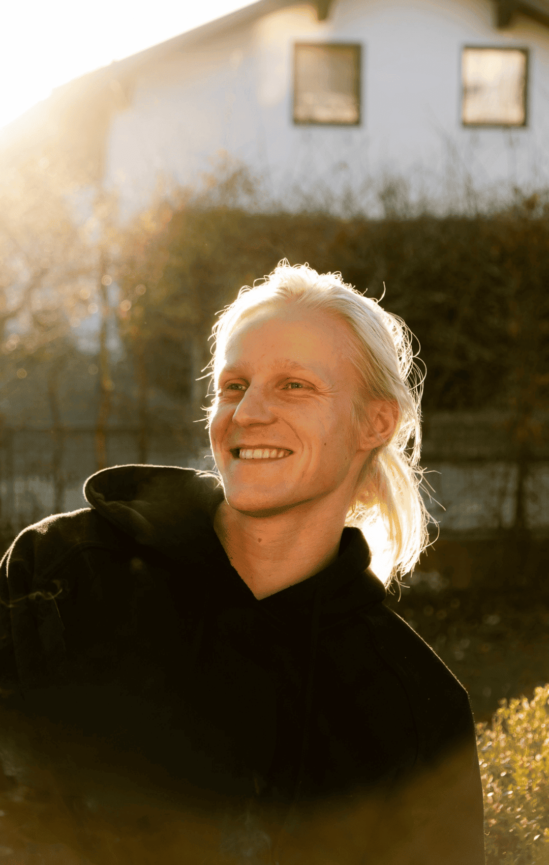 Lächelnder Mann mit langen, blonden Haaren im Freien bei Sonnenuntergang, trägt schwarze Jacke.