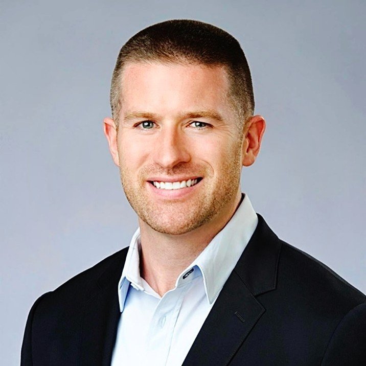 A smiling man in a white shirt and black suit jacket posing against a plain grey background.