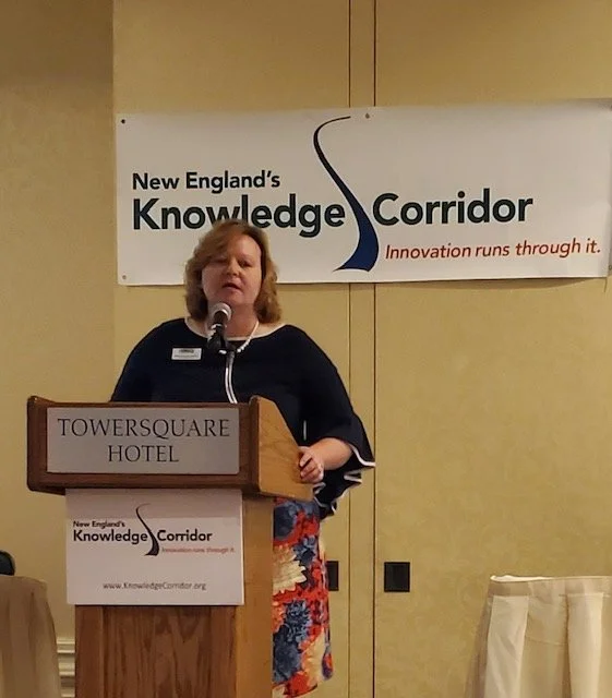 Woman speaking at podium during a conference, with a banner behind her reading 'New England's Knowledge Corridor' and a hotel sign reading 'Towersquare Hotel'.