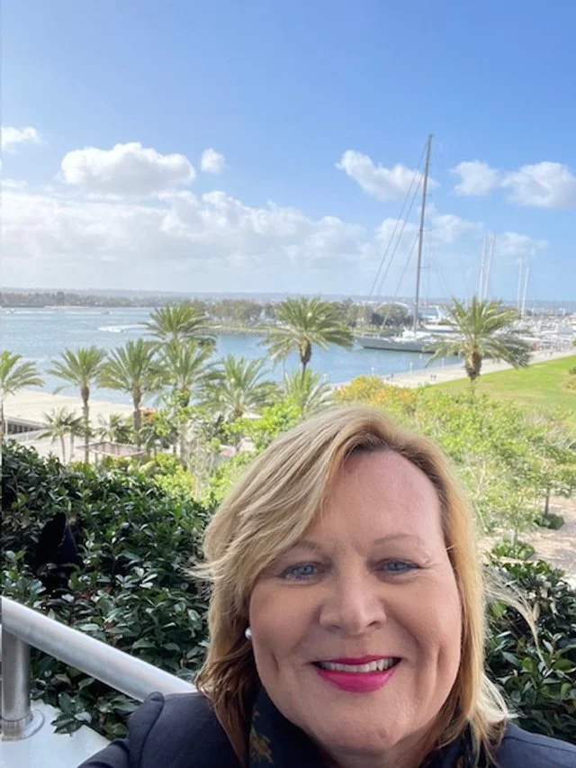 A woman smiling outdoors with a scenic waterfront view in the background, including palm trees, a marina with sailboats, and a partly cloudy sky.