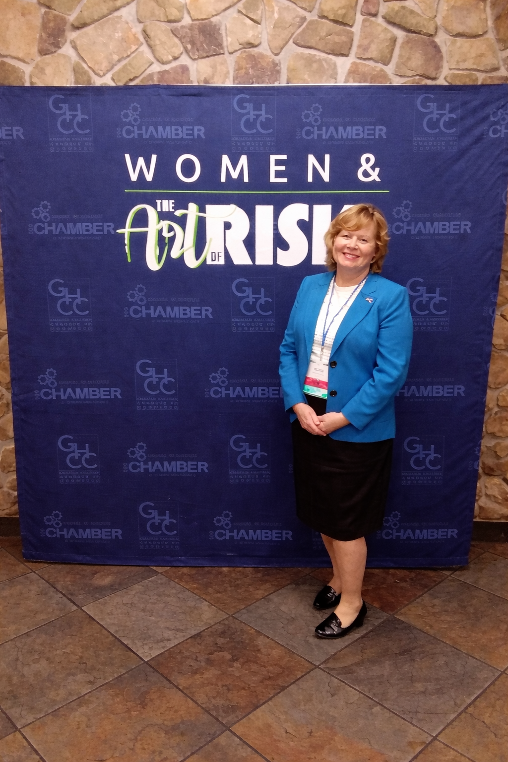 A woman in a blue blazer standing in front of a backdrop that reads 'Women & the Risk' at a conference or event.