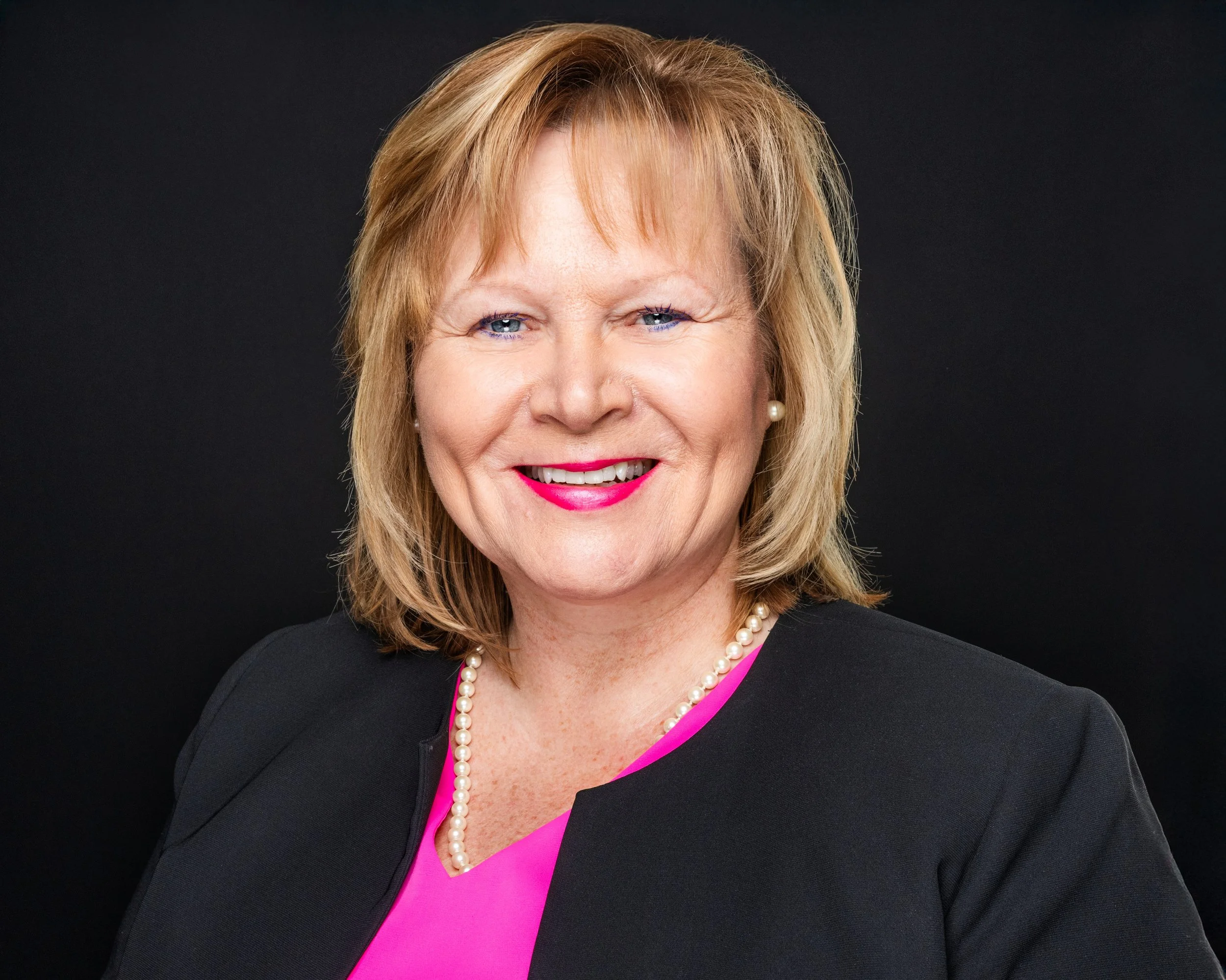 A woman with blonde hair, wearing a black jacket, pink blouse, pearl necklace, and pearl earrings, smiling against a black background.