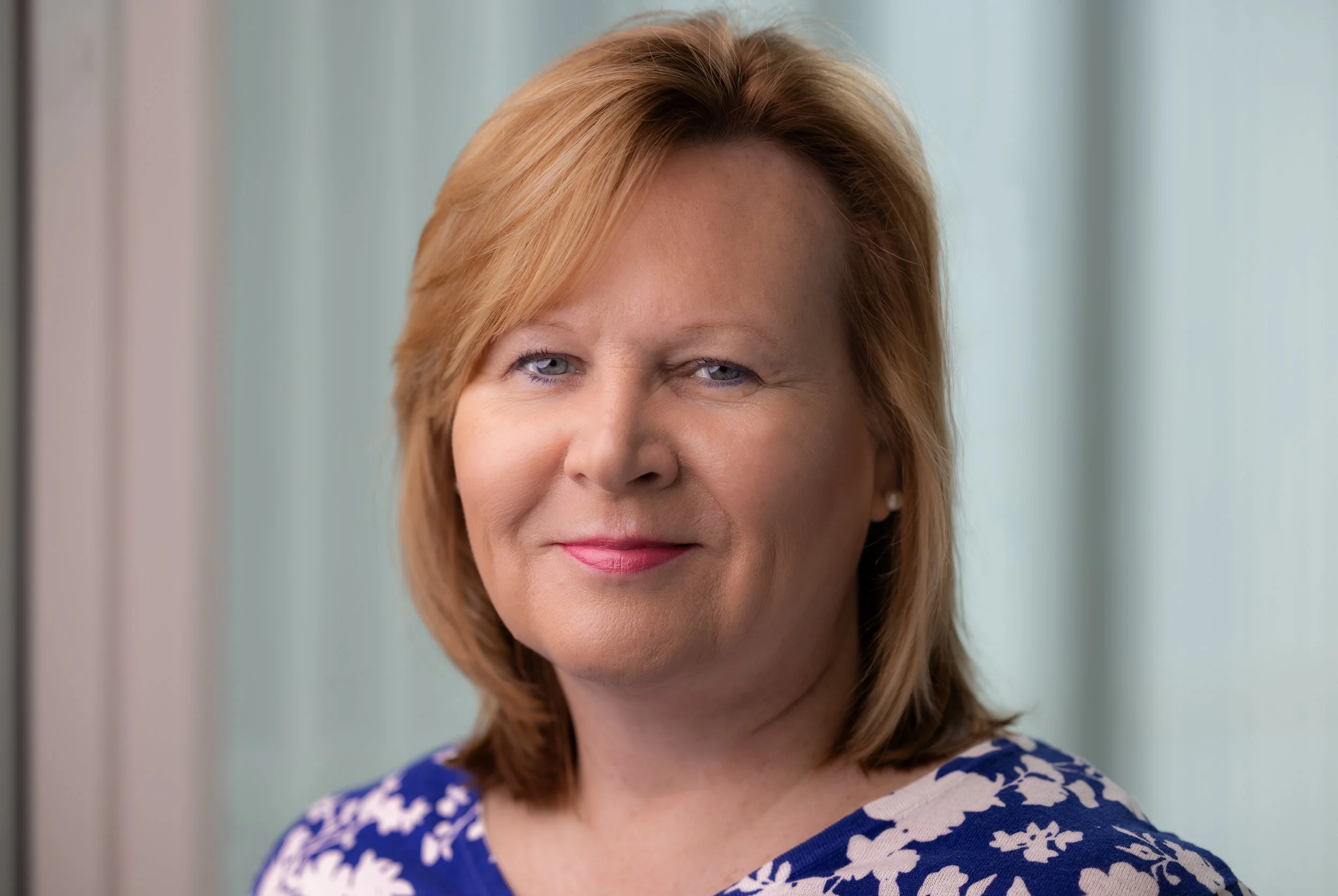 Portrait of a middle-aged woman with shoulder-length red hair, wearing a blue and white floral top, standing indoors near a window with light curtains.