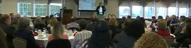 A conference room filled with attendees seated at tables, listening to a speaker at the front with a presentation screen behind them.