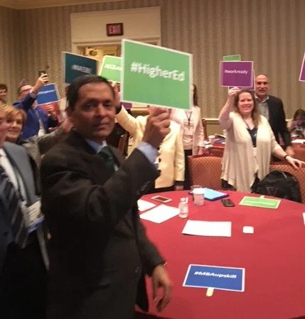 A group of people at a conference or gathering, holding signs with various hashtags like #highered, #workready, and others, with a man in the foreground holding a green sign.