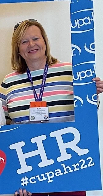 Smiling woman at a conference, holding a large blue frame with the HR hashtag and #cupahr22, wearing a striped shirt and conference badge.