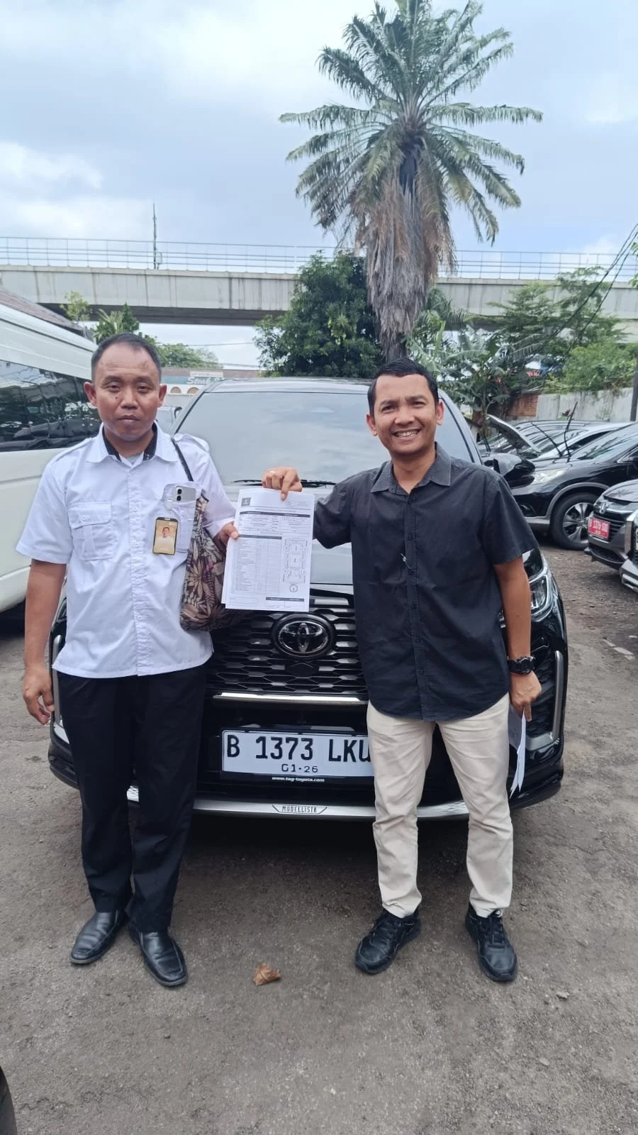 Two men standing in front of a black Toyota car, holding documents and smiling, with parked cars and a tall palm tree in the background.