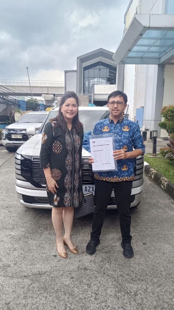 A woman and a man standing in front of a white vehicle, the man holding a document, outside a modern building with cloudy sky in the background.