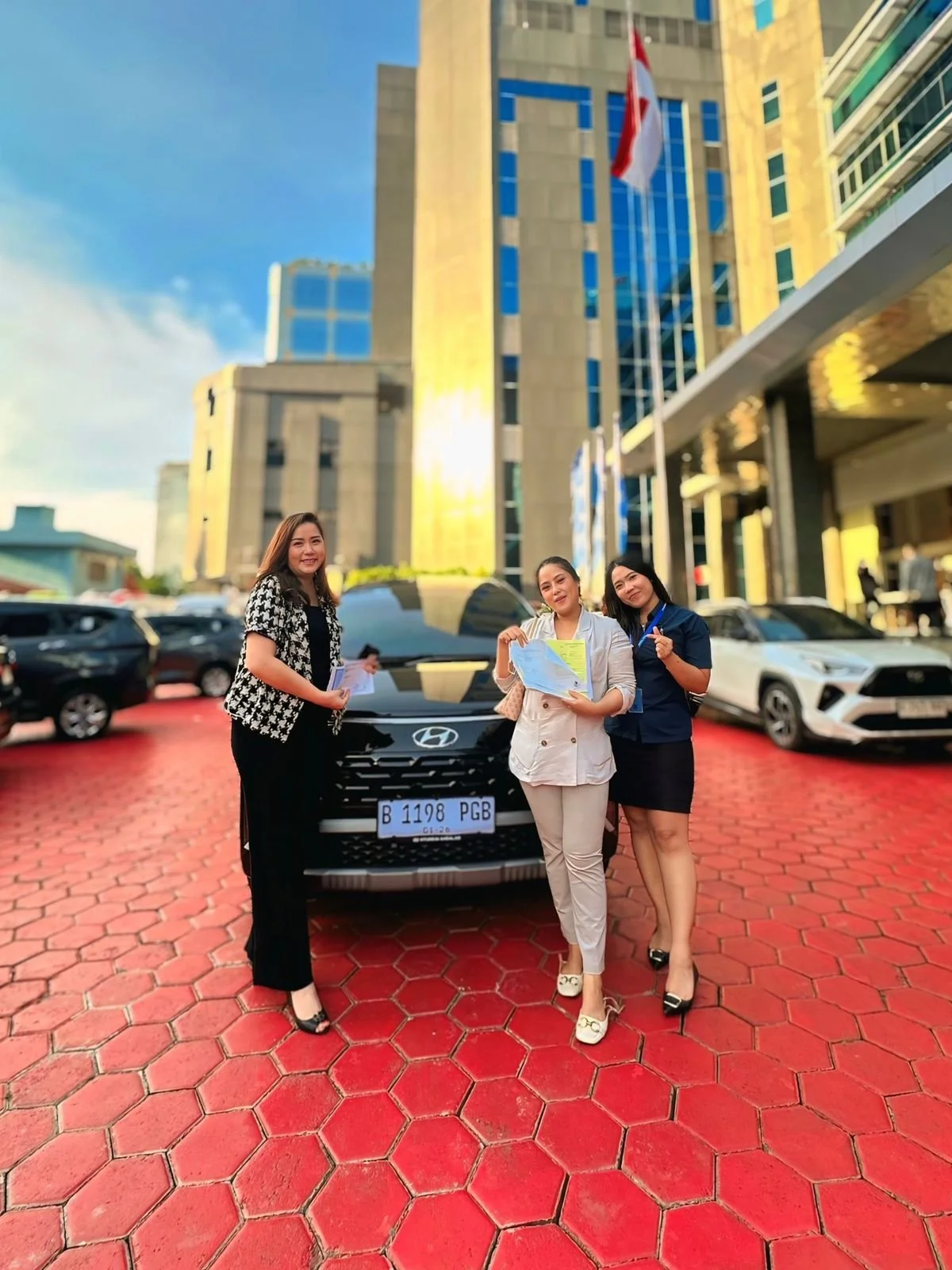 Three women standing in front of a black Hyundai car with a modern office building in the background, on a red hexagonal tile pavement.