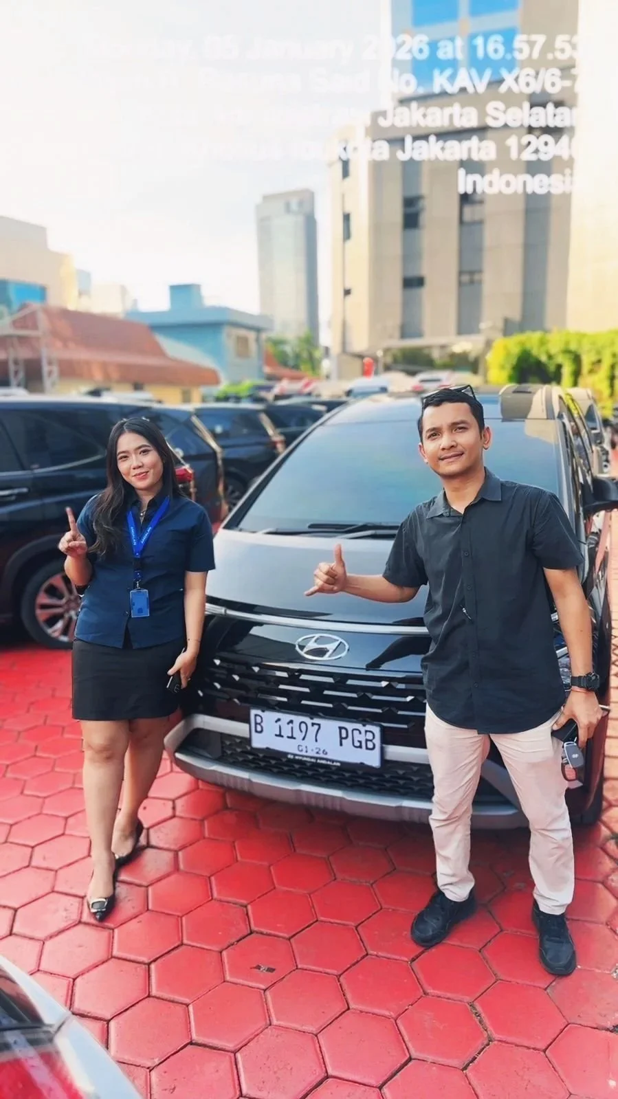 A man and woman standing in front of a black Hyundai car on a red paving stone lot in an urban area with tall buildings behind them. The woman is dressed in a dark blue top and black skirt, holding a phone, and making a peace sign. The man is wearing a black shirt and beige pants, holding a car key and making a shaka sign. Both are smiling at the camera.