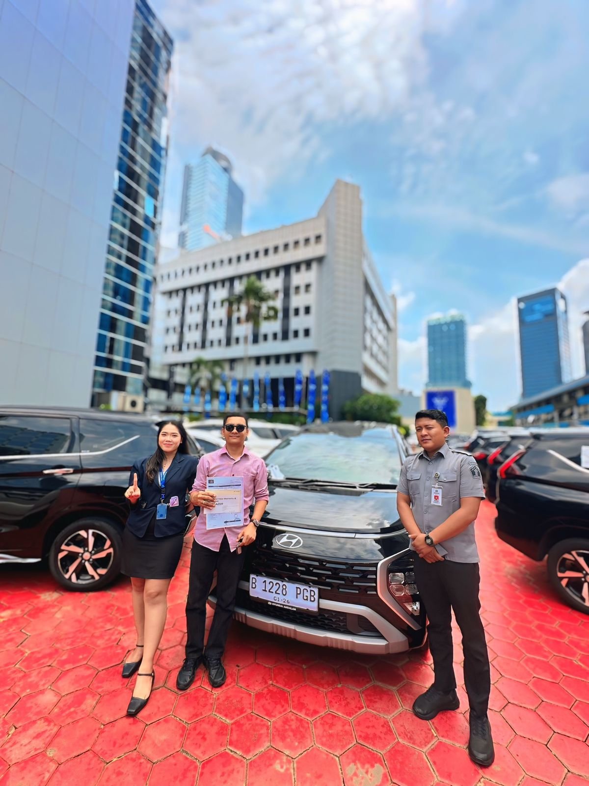 Three people standing in front of a black Hyundai car in a parking lot with tall buildings and a blue sky in the background.