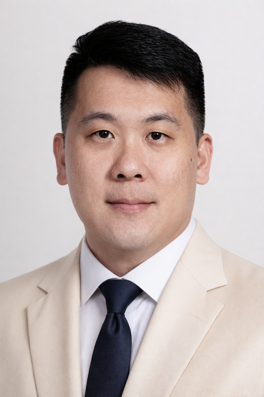 Headshot of a young Asian man wearing a beige suit, white shirt, and black tie against a plain white background.