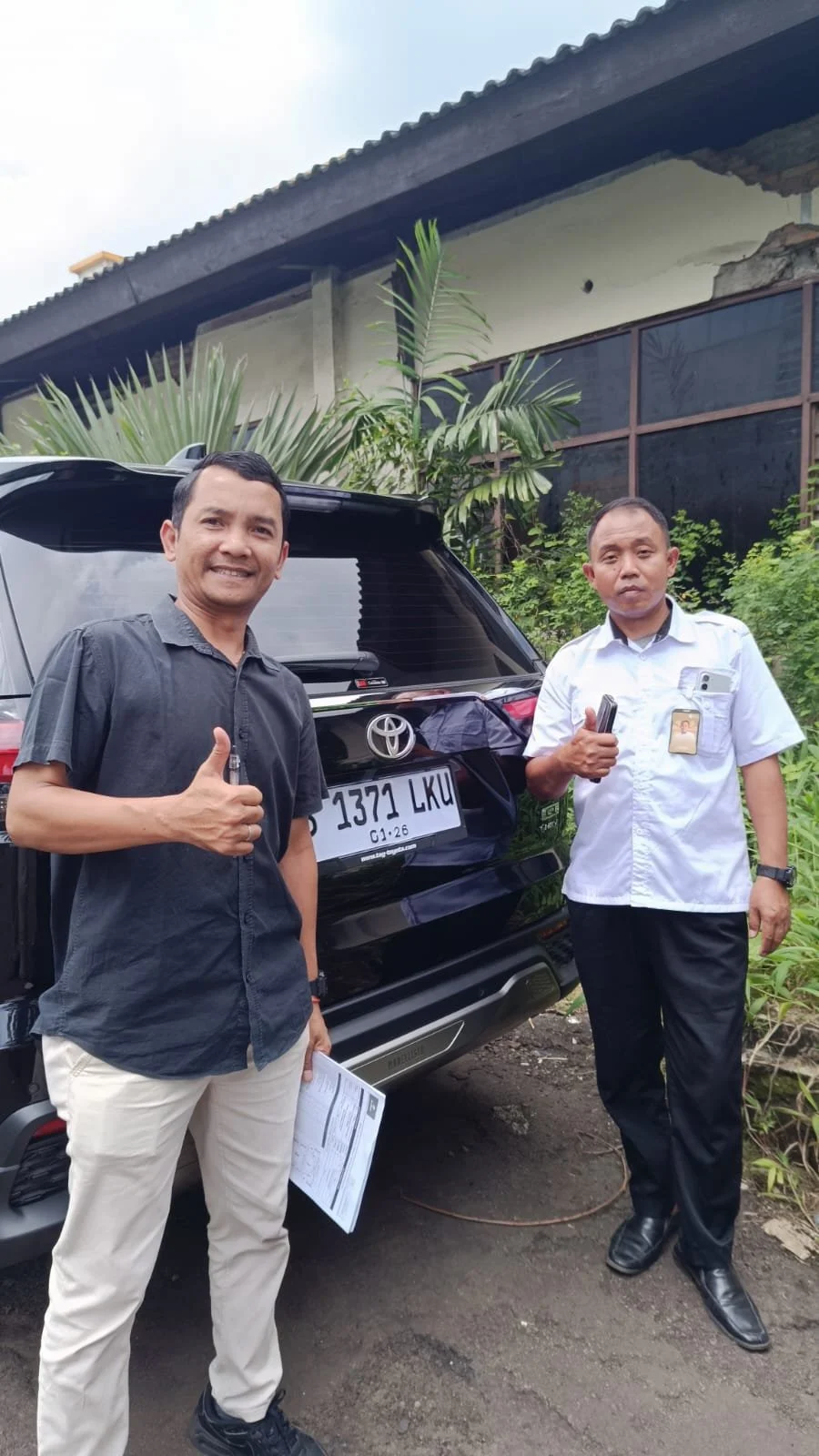 Two men standing in front of a black Toyota car with a license plate 1371 LKU, one giving a thumbs up and the other holding a phone, outdoors near a building and greenery.
