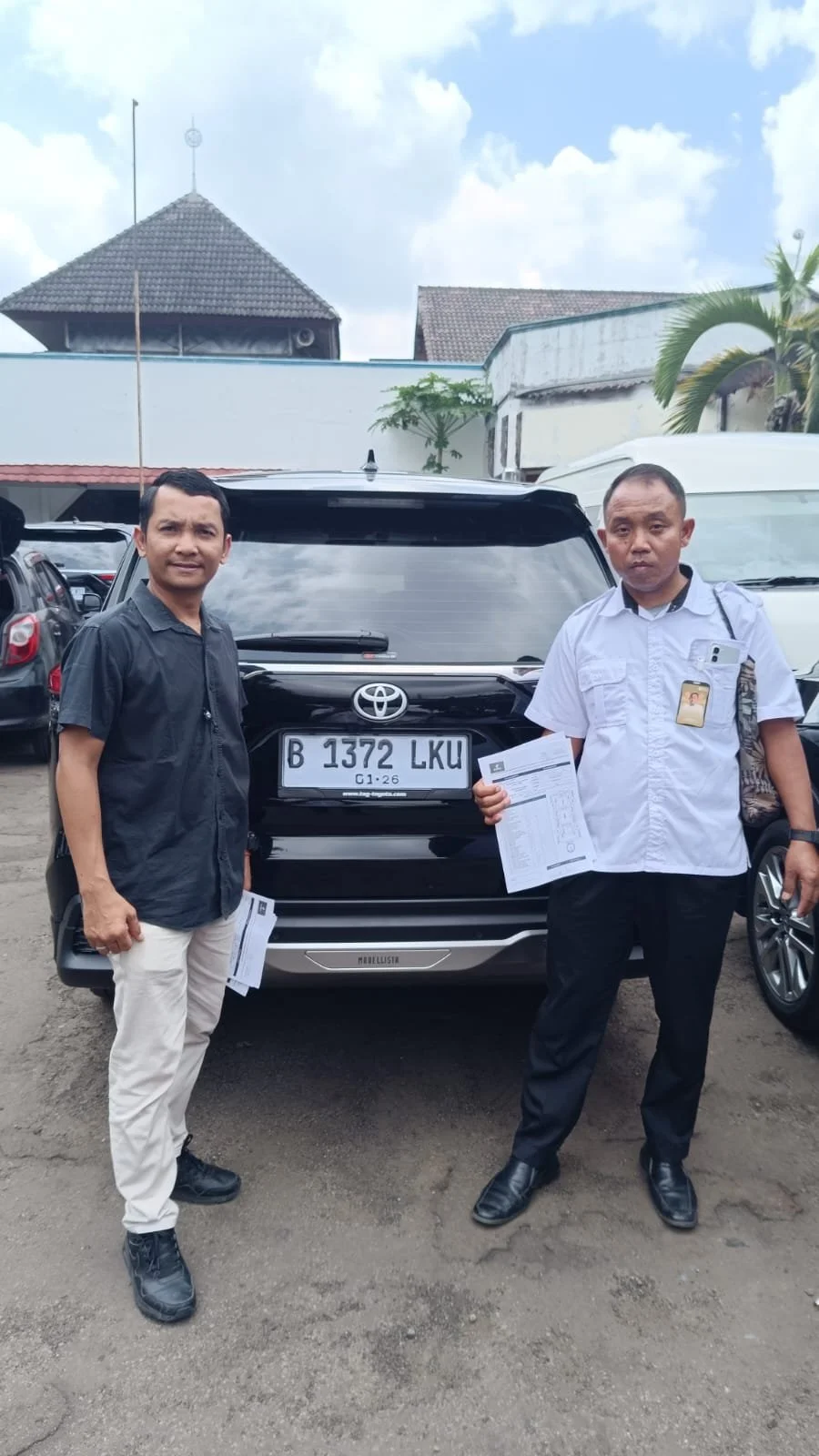 Two men standing in front of a black Toyota Land Cruiser with a license plate B 1372 LKU, holding documents, in a parking lot with multiple cars and buildings in the background.