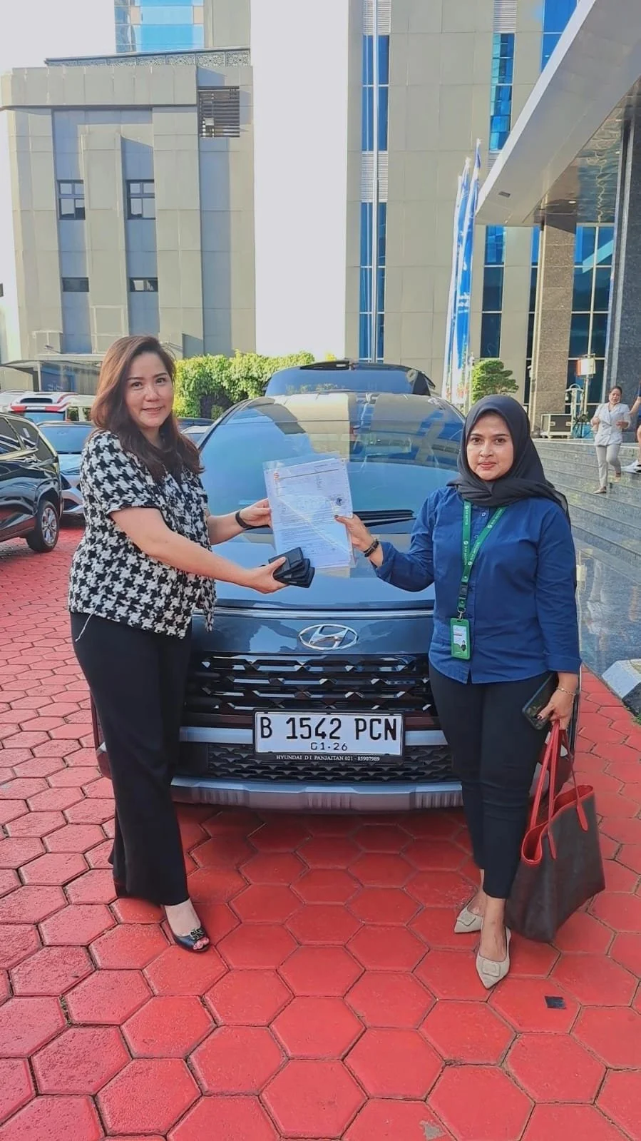 Two women standing in front of a Hyundai car, holding documents and shaking hands, outside a modern office building.