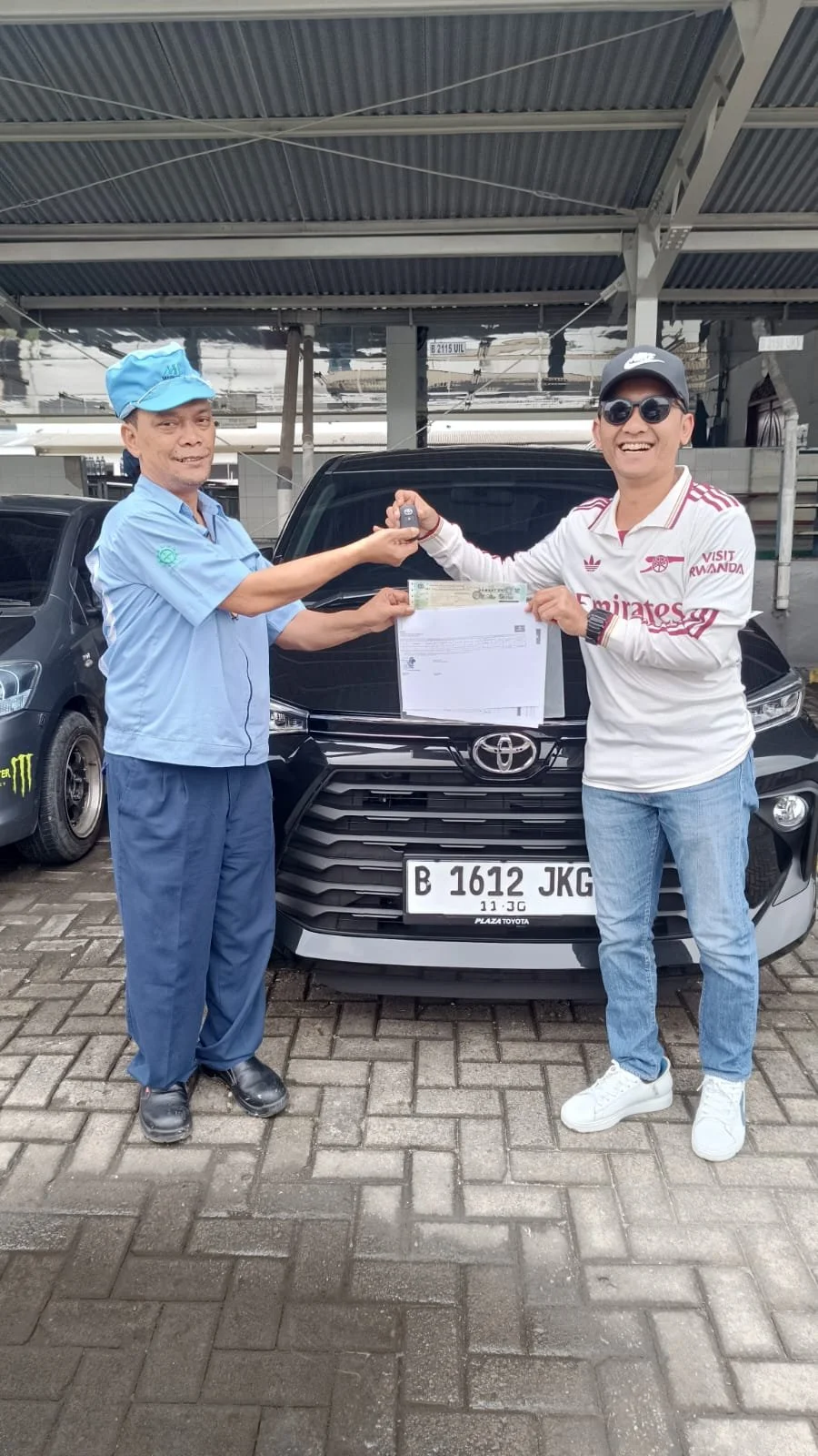 A man in a white shirt and sunglasses is receiving car keys and documents from a man in a blue uniform at a parking lot, with a black Toyota car in the background.