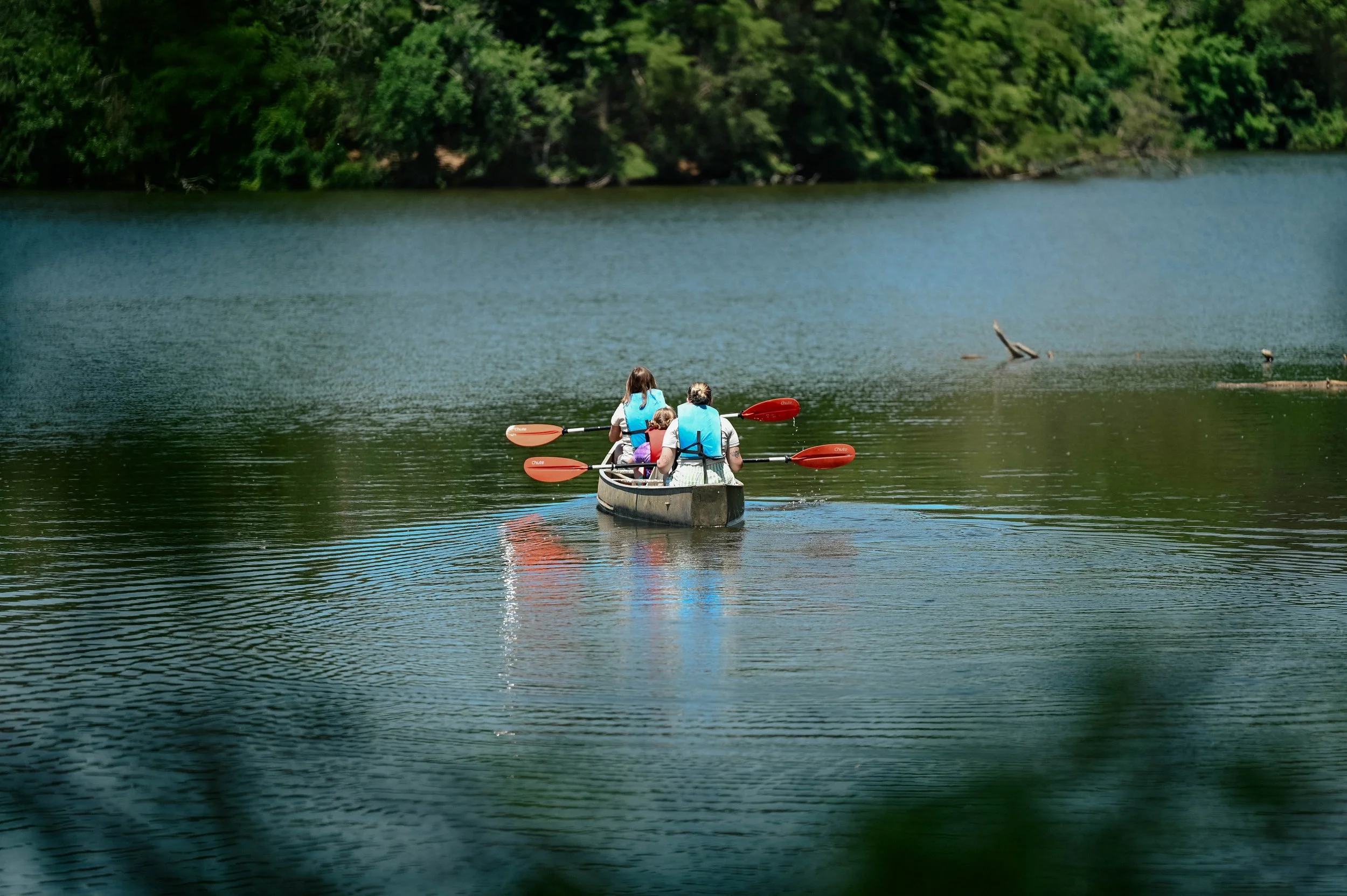 Two female camp counselors in a canoe with a female camper on an open lake with some trees in the background.