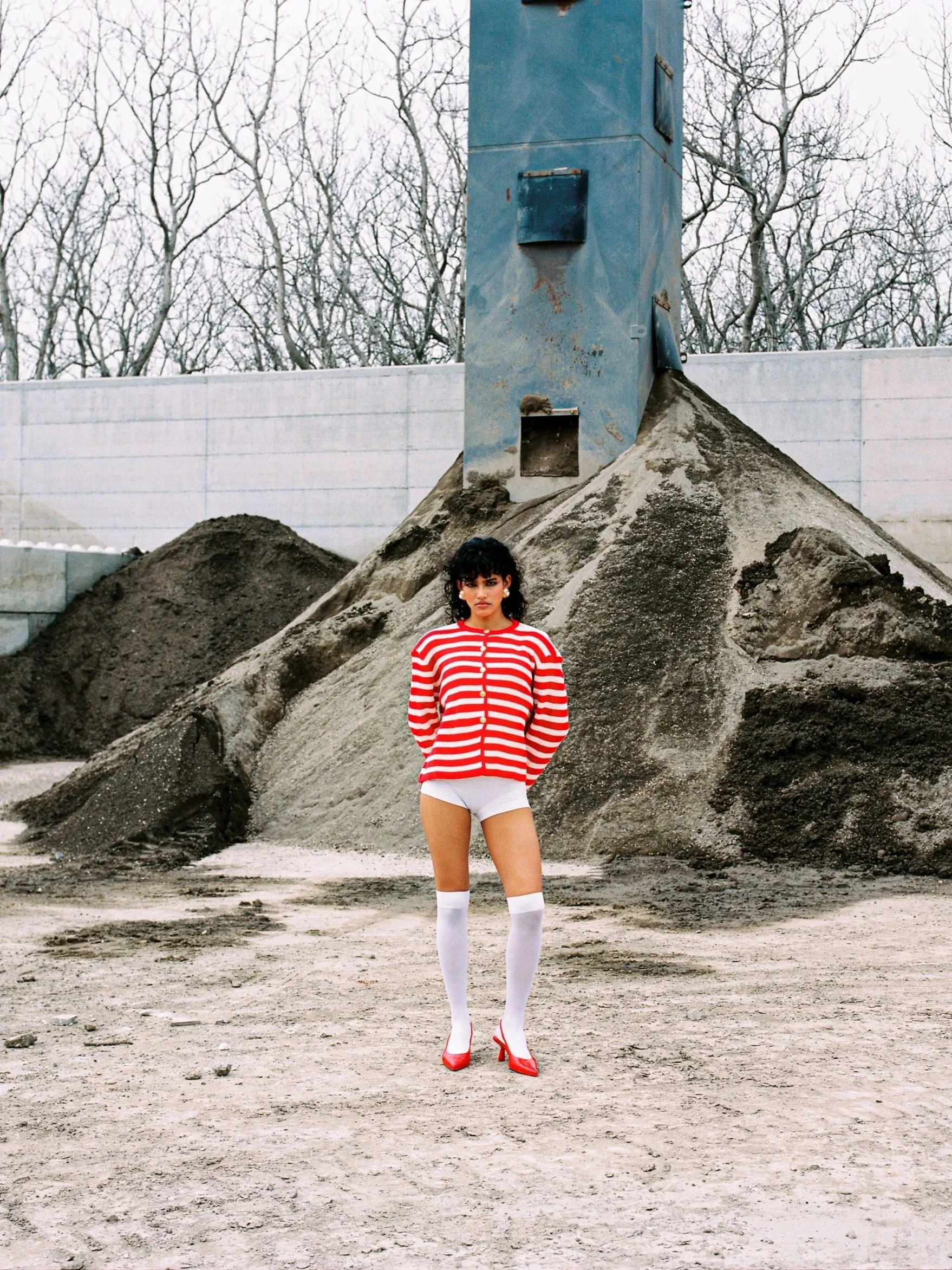 A woman in a red and white striped jacket, white shorts, thigh-high socks, and red heels standing in front of a construction site with dirt piles and an industrial blue structure with leafless trees in the background.