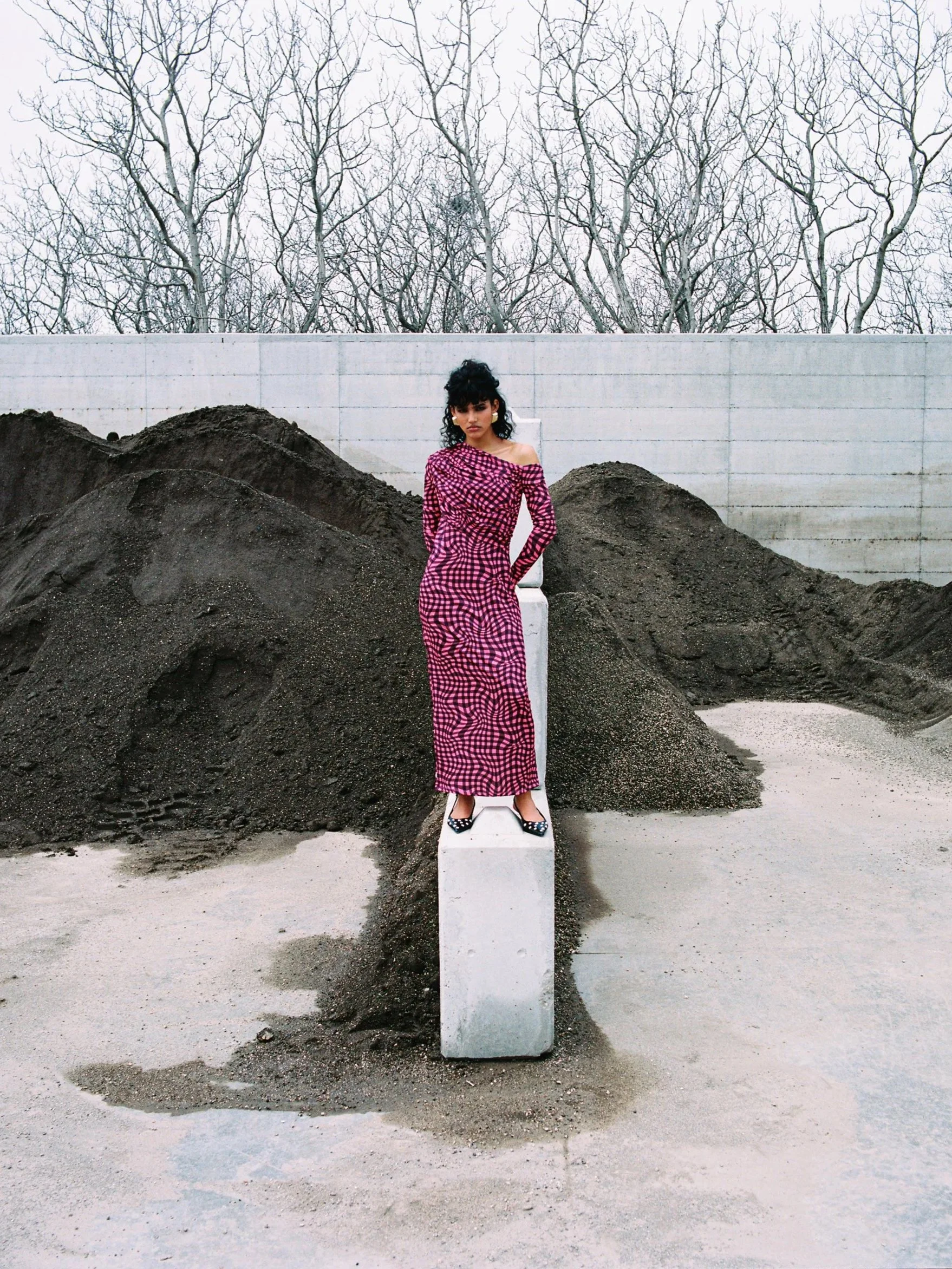 A woman wearing a pink and black checkered off-shoulder dress, standing on a white concrete block amidst piles of dirt, with a background of leafless trees and a gray wall.