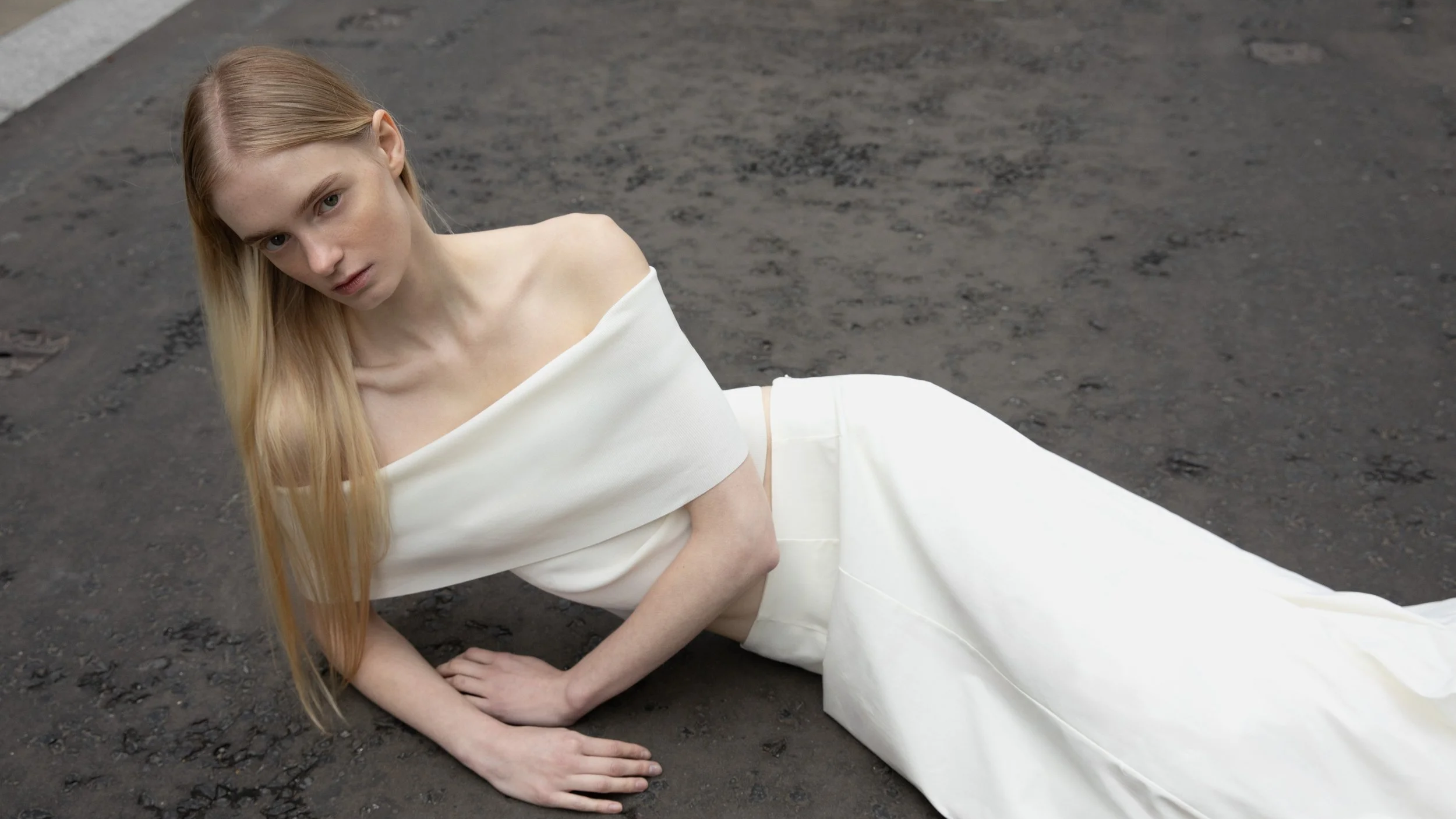 A young woman with long blonde hair lying on a rough, dark asphalt surface, wearing a white off-shoulder dress.