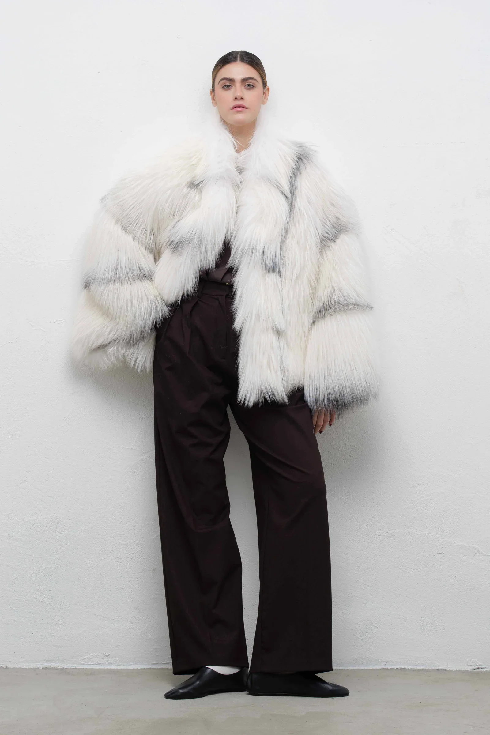 Woman wearing a white fur coat, dark pants, and black shoes standing against a plain white wall