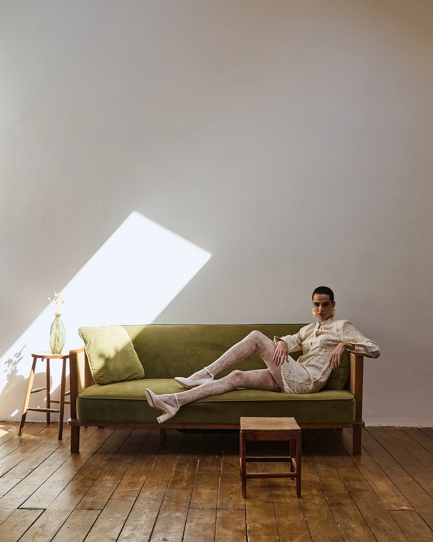 Woman reclining on a green vintage sofa in a minimalist room with wooden floor, small side table with a vase, and a wall with a geometric shadow pattern.