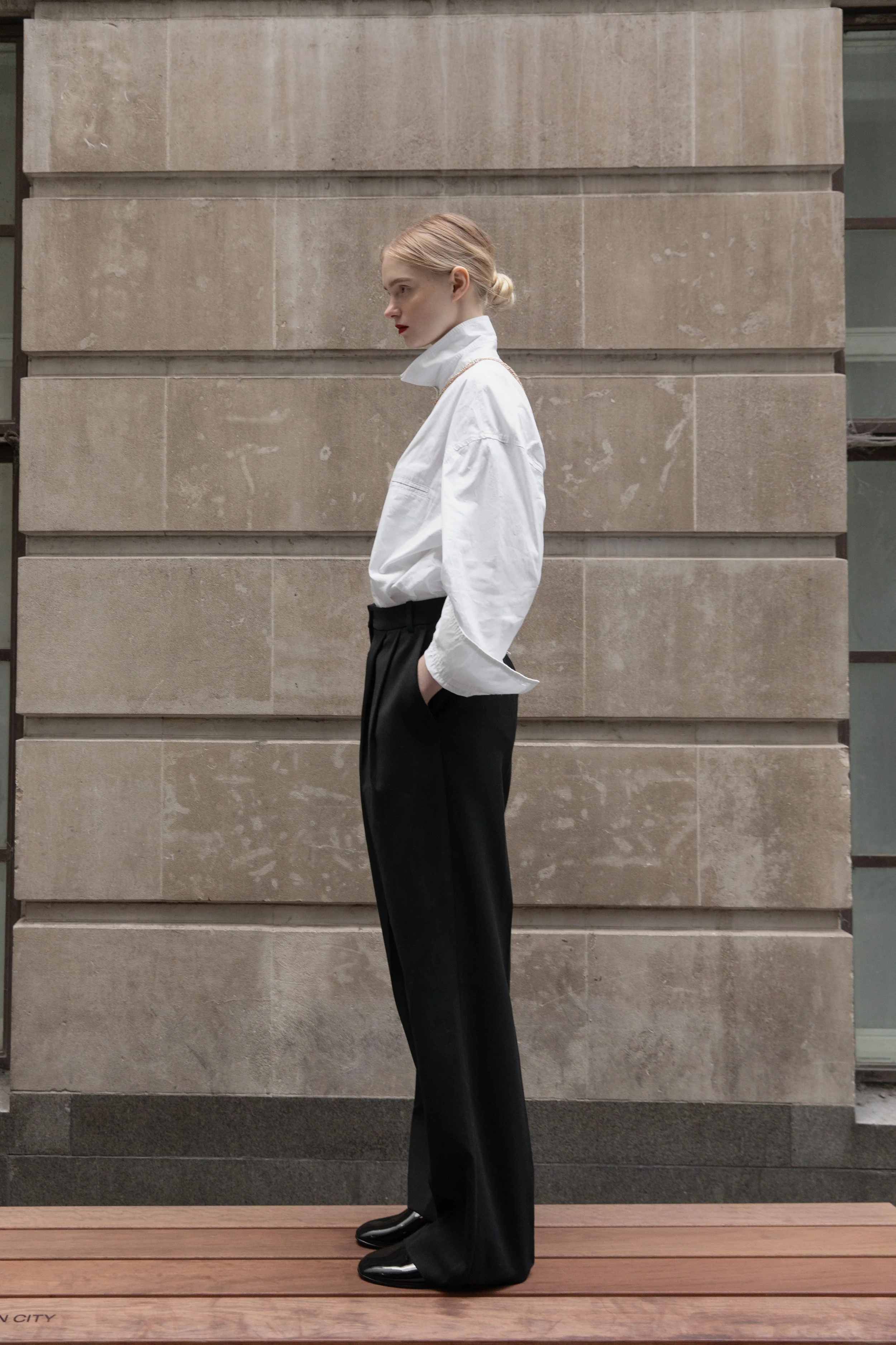 A woman with blonde hair tied in a bun, wearing a white oversized shirt with a high collar, black wide-leg trousers, and black shiny shoes, standing in profile against a beige stone wall on a wooden platform.