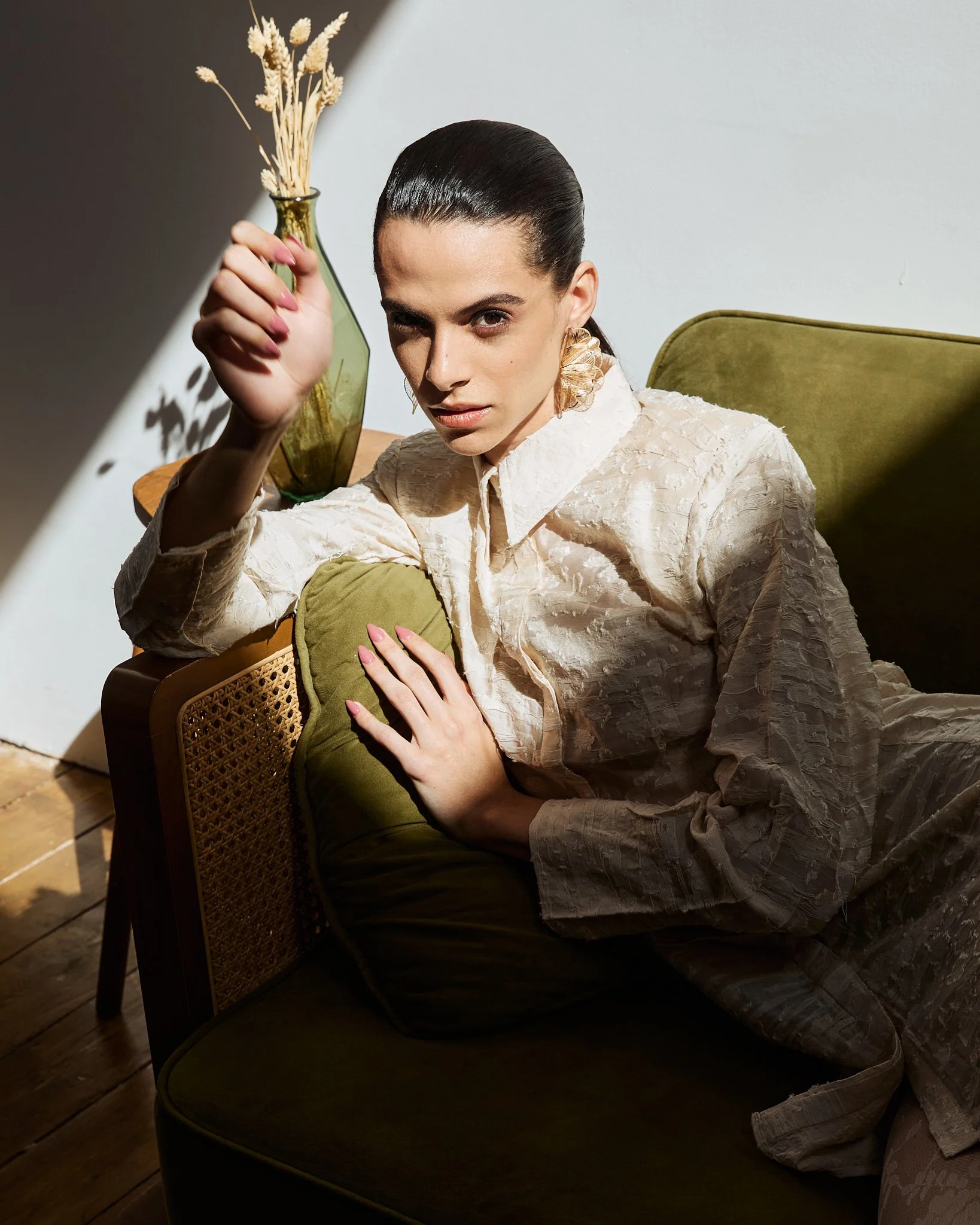 A woman with dark hair tied back, wearing a light-colored textured dress or coat, is reclining on a green velvet chair. She holds a small green pillow with her left hand and raises a green glass vase with dried flowers in her right hand. She is looki