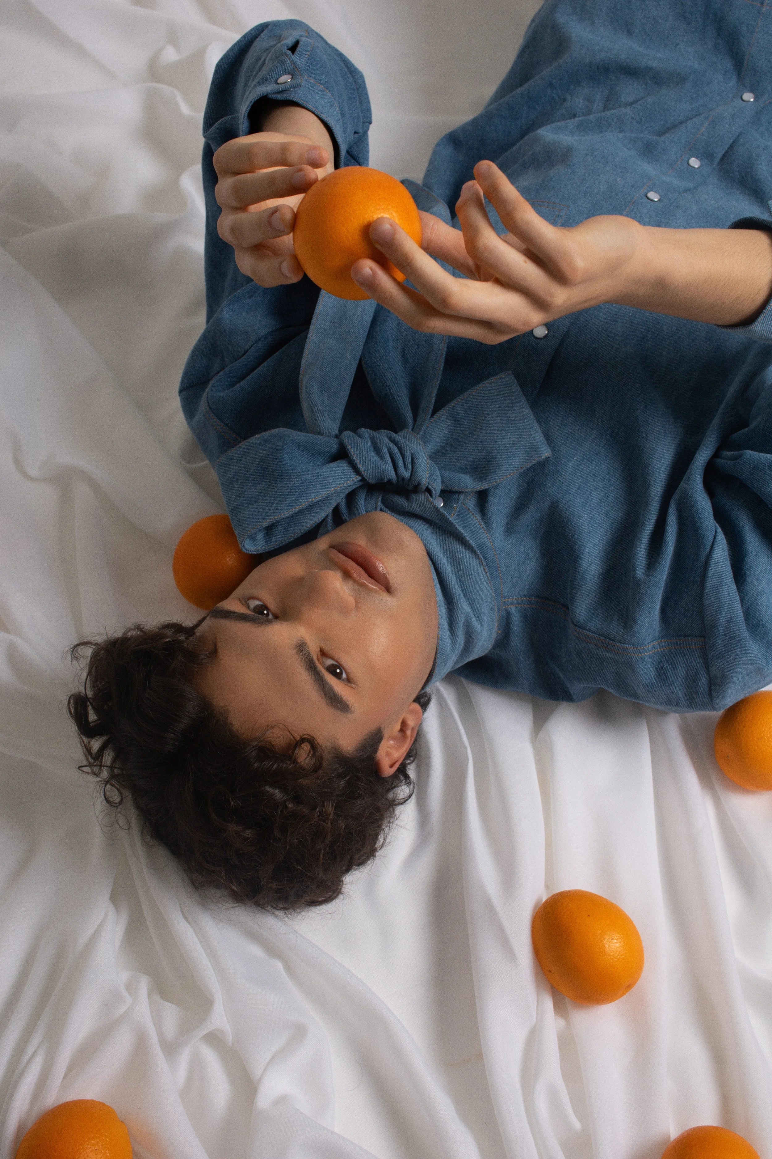 A person lying on a white bed surrounded by oranges, holding one orange in their hand, wearing a blue denim shirt with a tie at the collar.