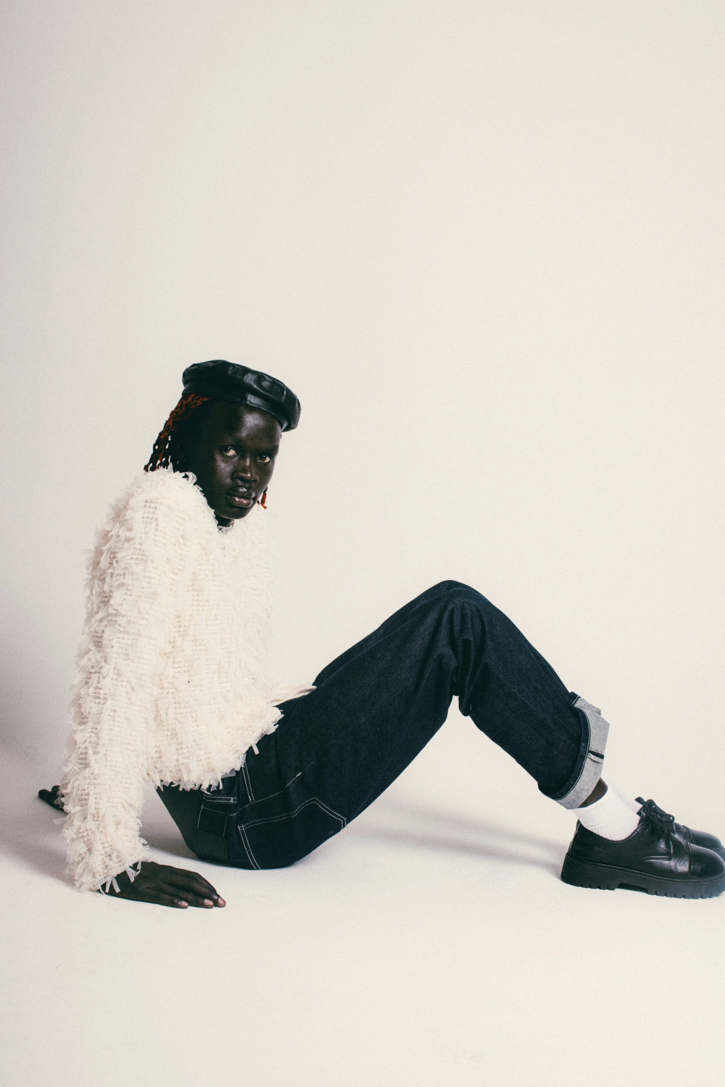 A person with dark skin and orange dreadlocks sitting on the floor against a plain white background. They are wearing a black beret, a textured white sweater, cuffed dark jeans, white socks, and black shoes. The person is looking directly at the came