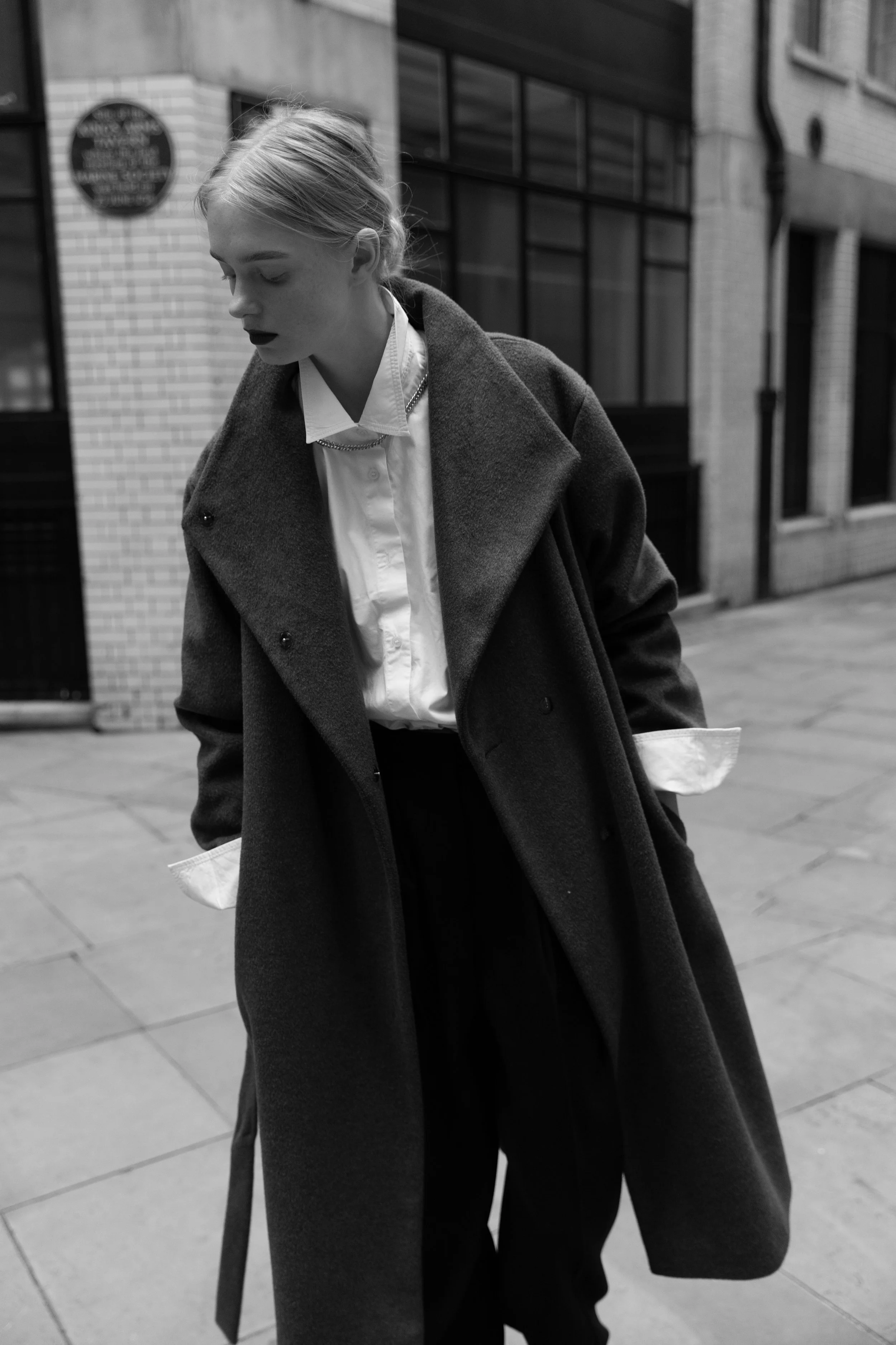 A woman with light hair, wearing a dark overcoat, white shirt with rolled-up sleeves, and dark pants, stands on a city sidewalk with brick and glass buildings in the background.