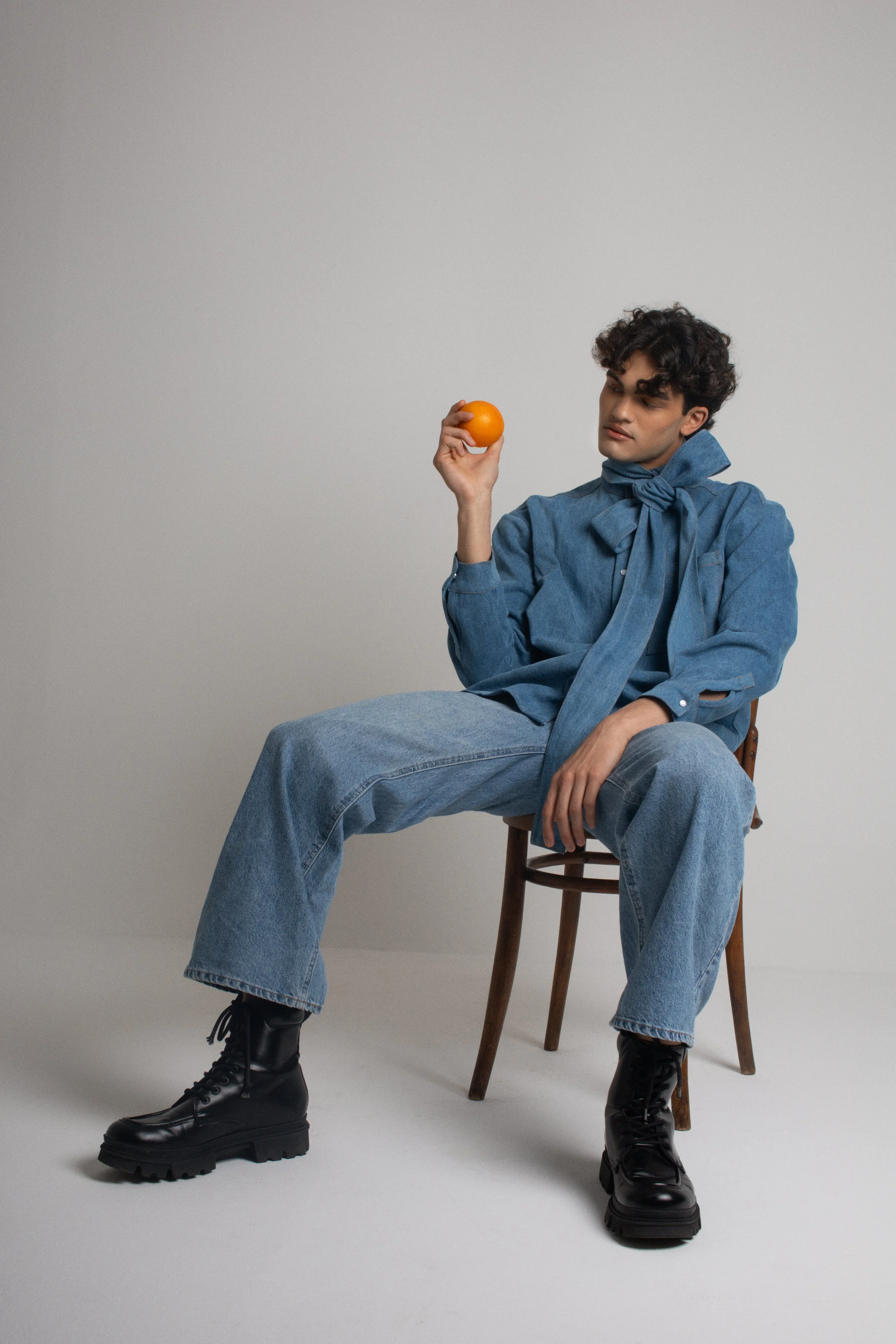 A young person with curly dark hair sitting on a wooden chair, wearing a denim outfit with a matching scarf, holding an orange in their right hand, and looking at it thoughtfully against a plain light gray background.