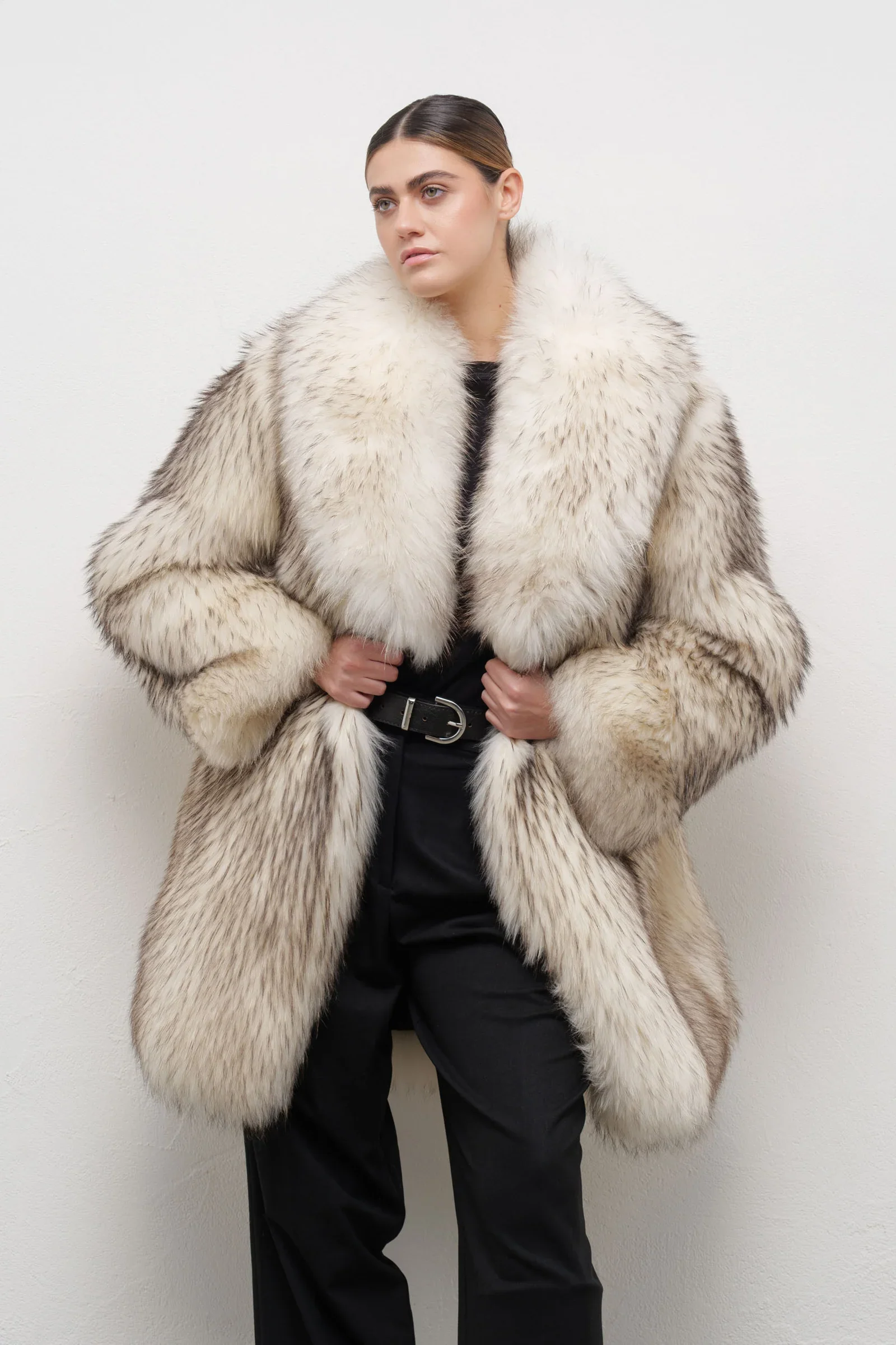 Woman wearing a luxurious cream-colored fur coat with black pants, standing against a plain white wall.