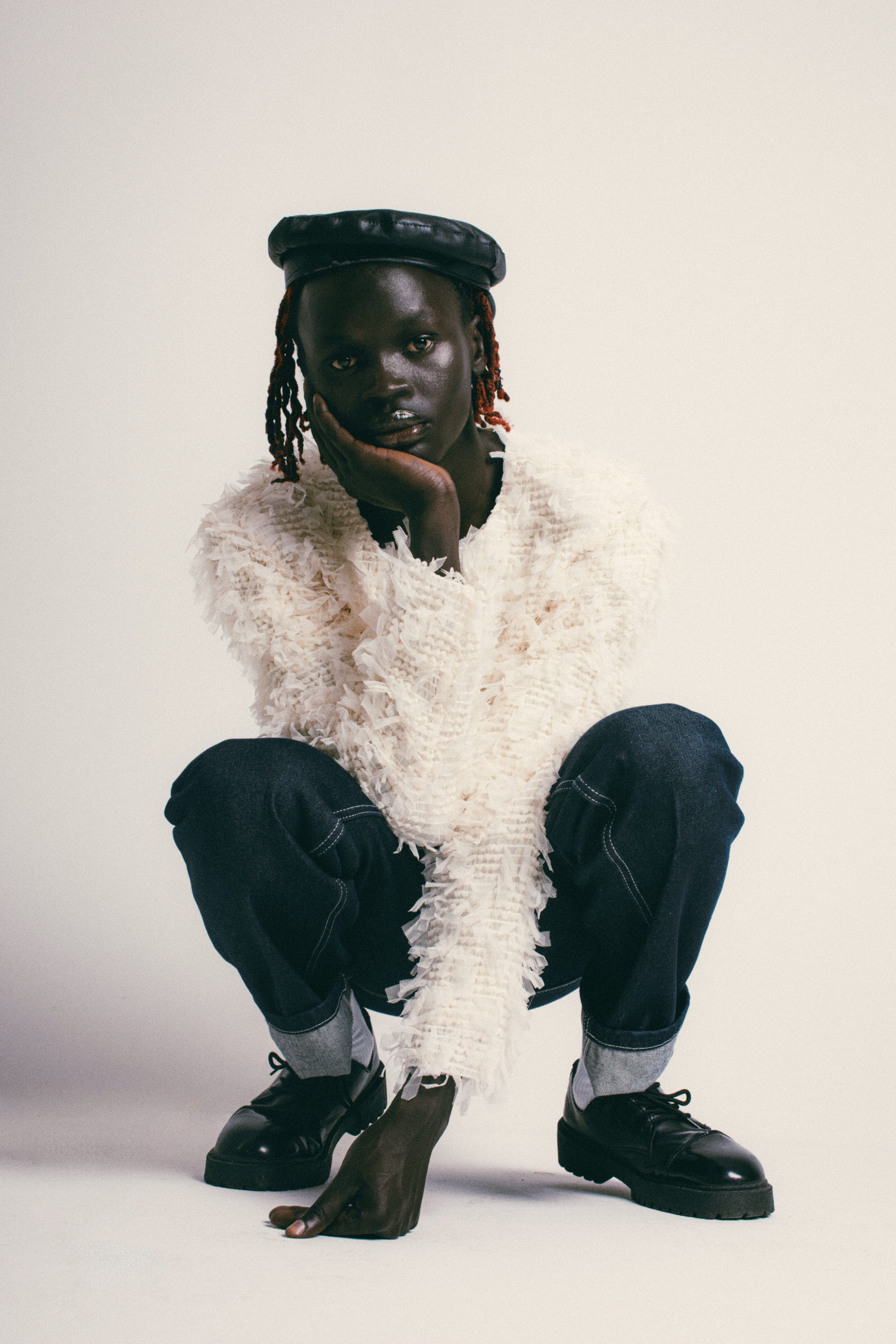 A person with dark skin and red-dyed hair wearing a black leather cap, a fluffy white textured jacket, dark jeans, and black shoes, squatting on the floor with their hand resting on their face against a plain light background.