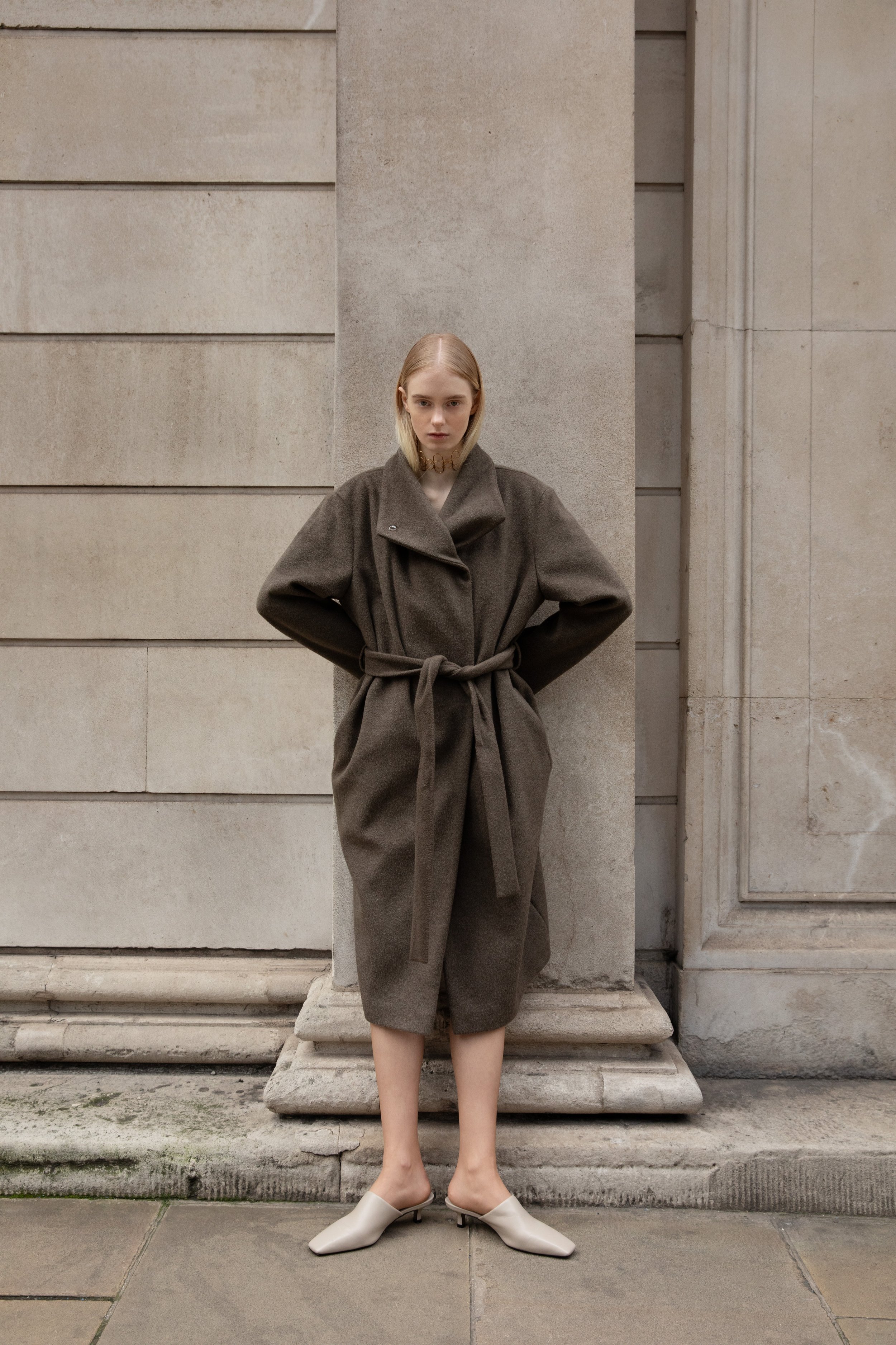 A woman standing against a stone wall in a beige coat with a belt, wearing beige heeled shoes.