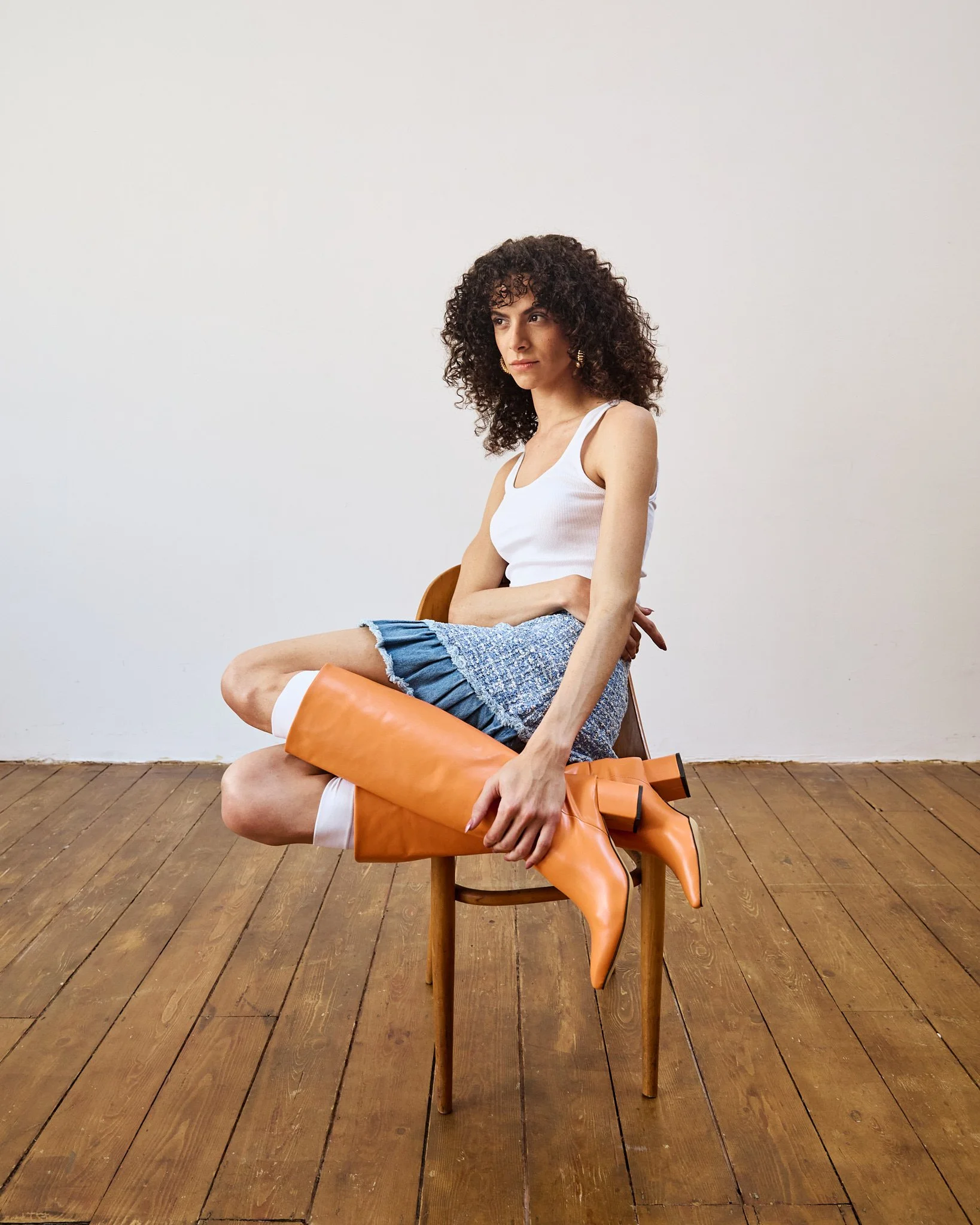 A woman with curly hair wearing a white tank top, a blue skirt, and bright orange knee-high boots, sitting on a wooden chair against a plain white wall and wooden floor, with her legs crossed and a contemplative expression.