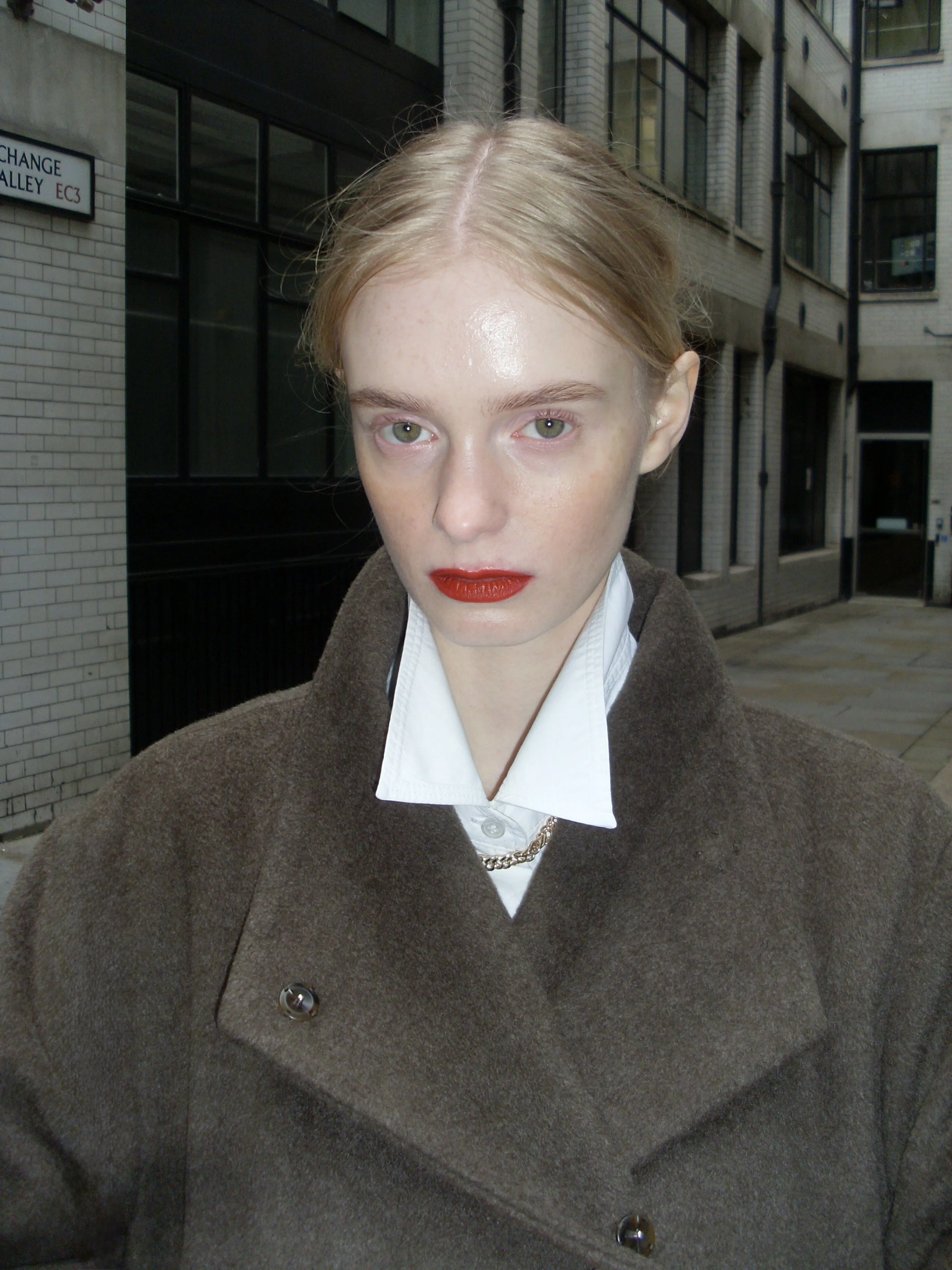 Close-up of a woman with red lipstick, wearing a white collared shirt and brown coat, standing outdoors in an urban area with brick buildings.