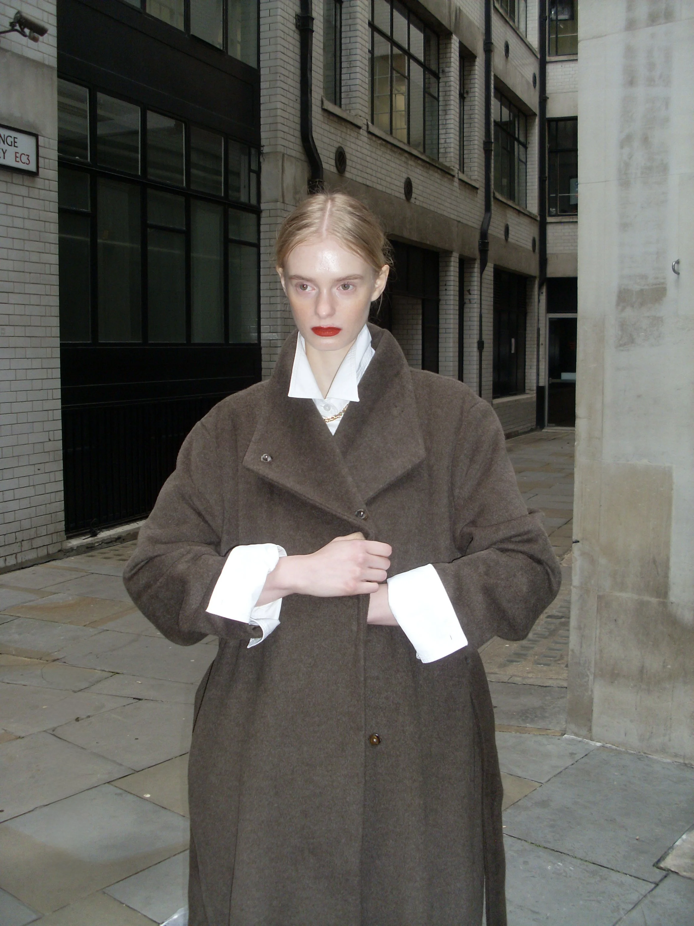 A woman with red lipstick and pale skin, dressed in a brown coat and white shirt, stands on a city sidewalk with modern brick and concrete buildings behind her.