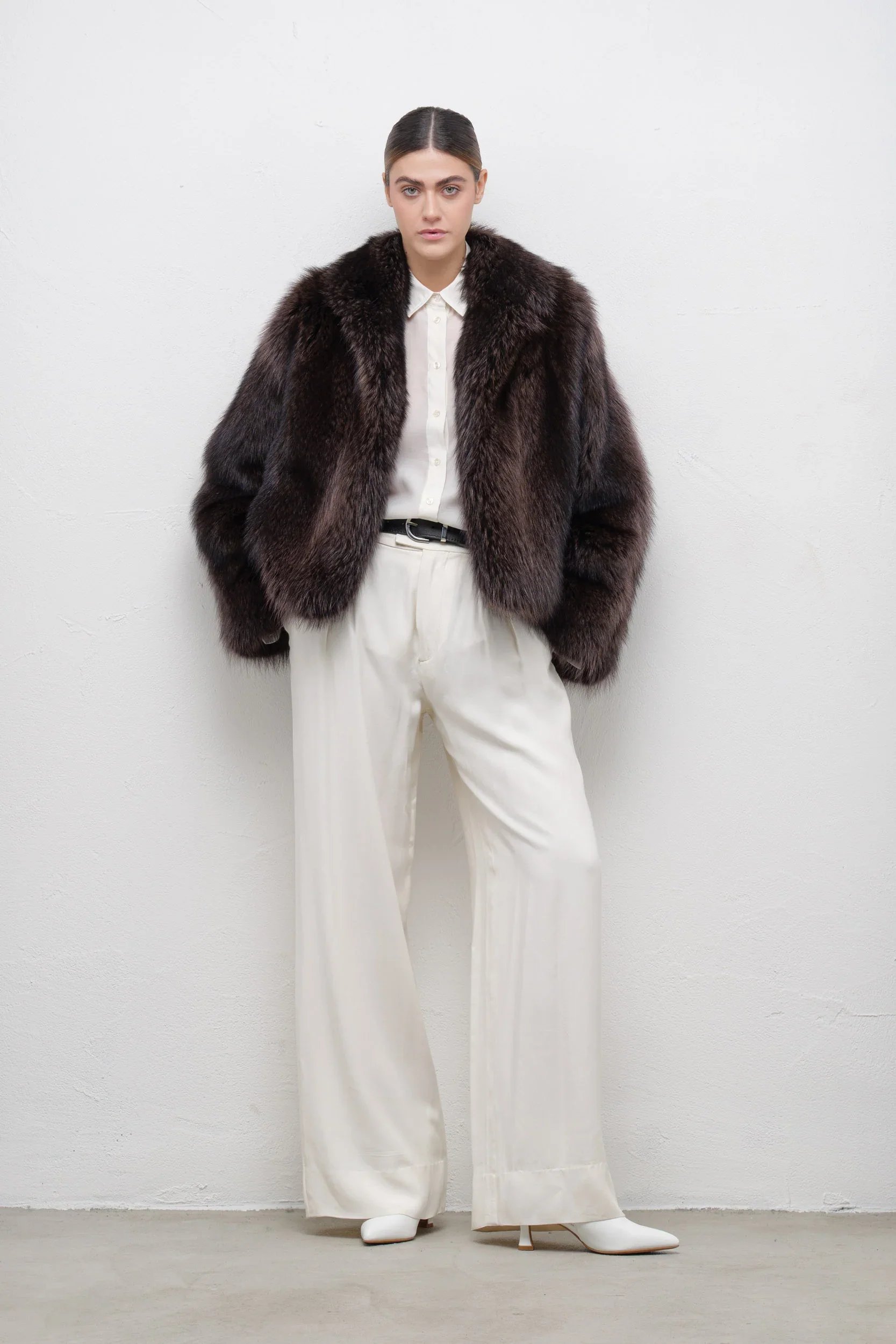 A woman with dark hair pulled back wearing a dark fur coat, white shirt, white wide-leg trousers, and white shoes stands against a plain white wall.