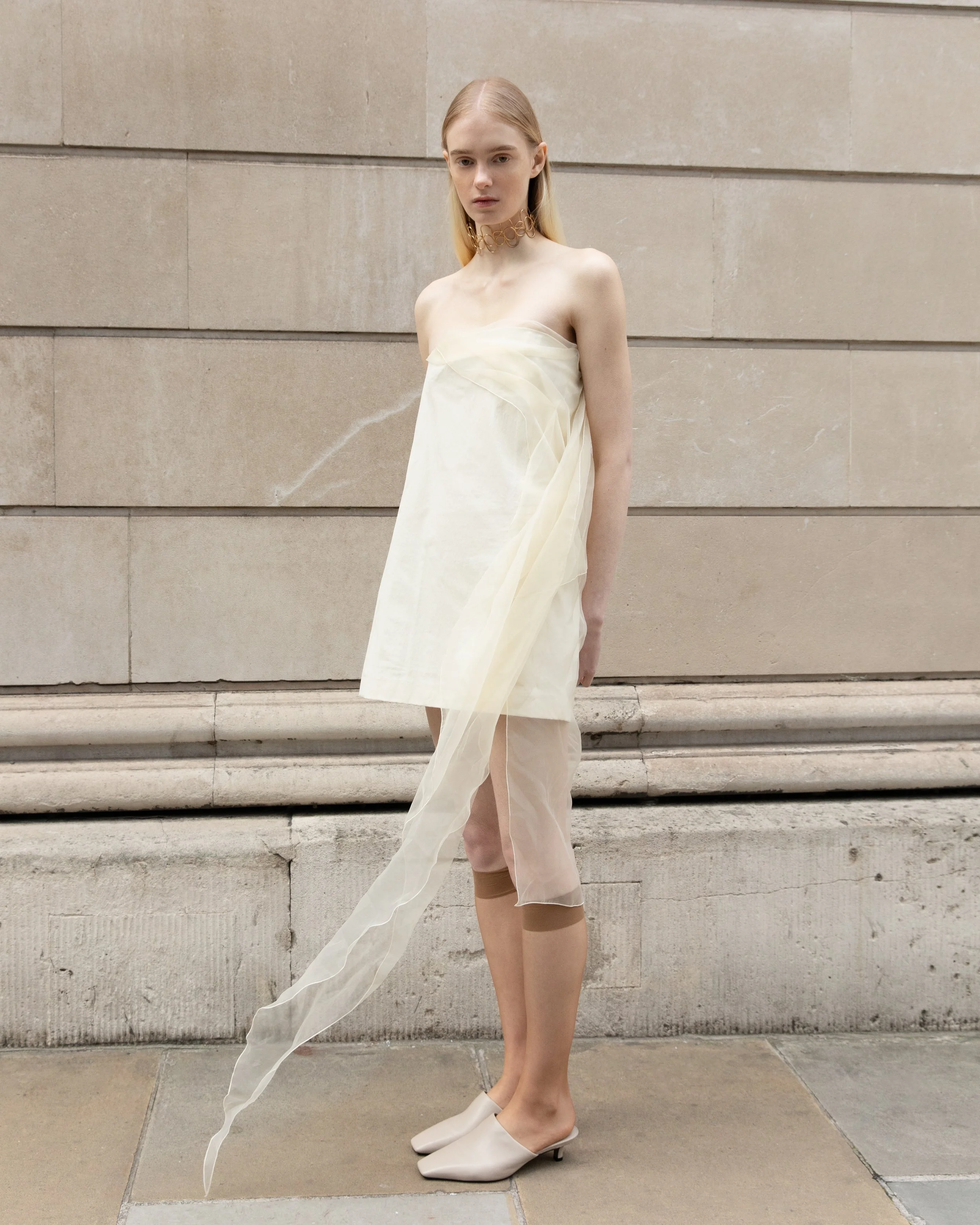 A young woman with blonde hair and pale skin standing against a stone wall overlooking a city street. She wears a strapless cream-colored dress, a gold choker necklace, and beige heels.