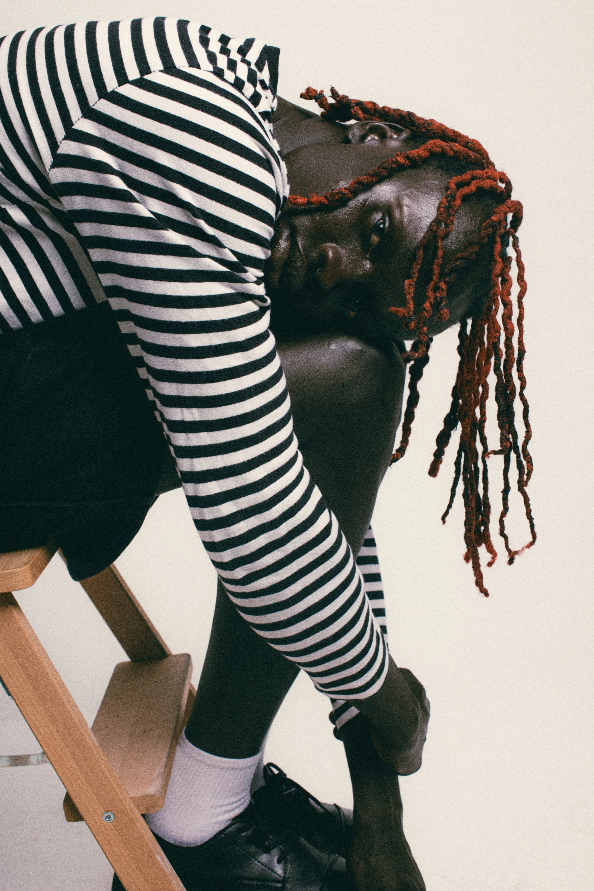 A person with dark skin and red dreadlocks lying on a dark chair, wearing a black and white striped shirt and black pants, with a neutral background.