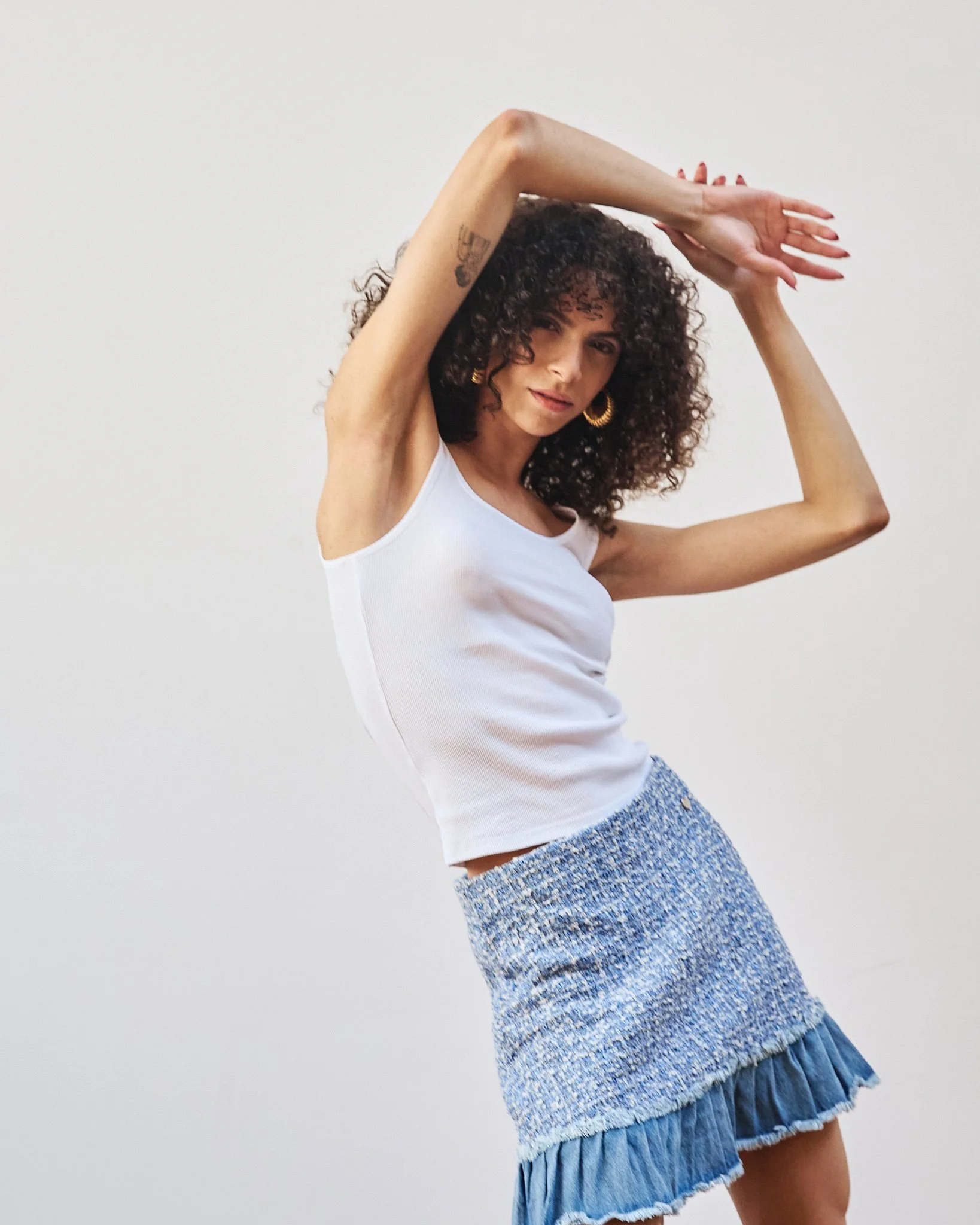 A woman with curly hair, wearing a white sleeveless top and a blue textured skirt with ruffled hem, with her arms raised and crossing above her head.
