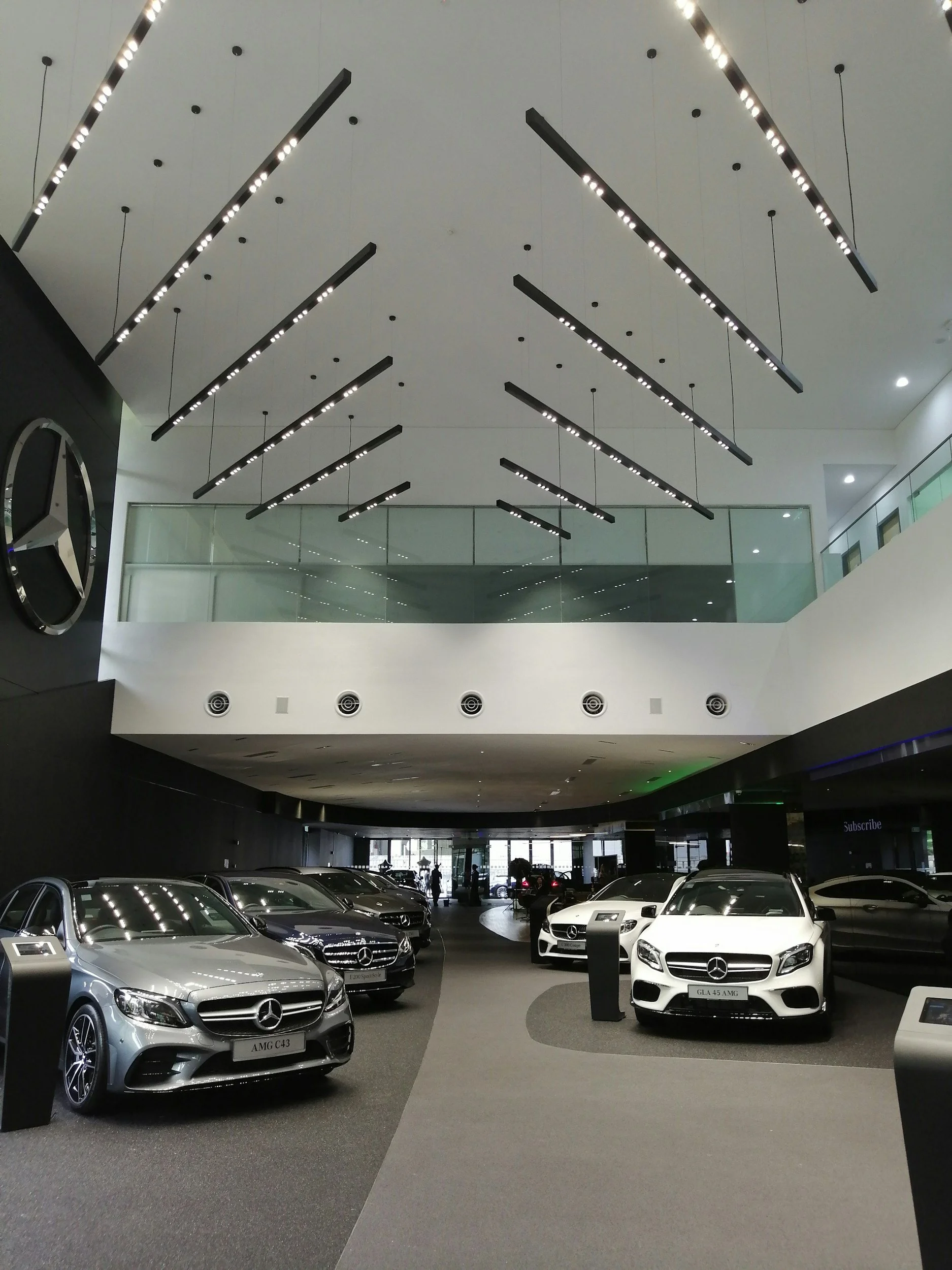 Showroom with Mercedes-Benz cars on display, including models AMG C43 and GLA 45 AMG, inside a modern dealership with black and white interior design, large ceiling lights, and a glass balcony.