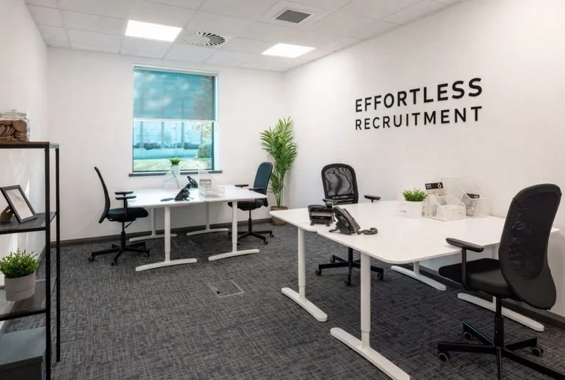 Modern office with white walls, black chairs, and white desks, featuring a sign on the wall that says 'Effortless Recruitment' and a window with a view outside.